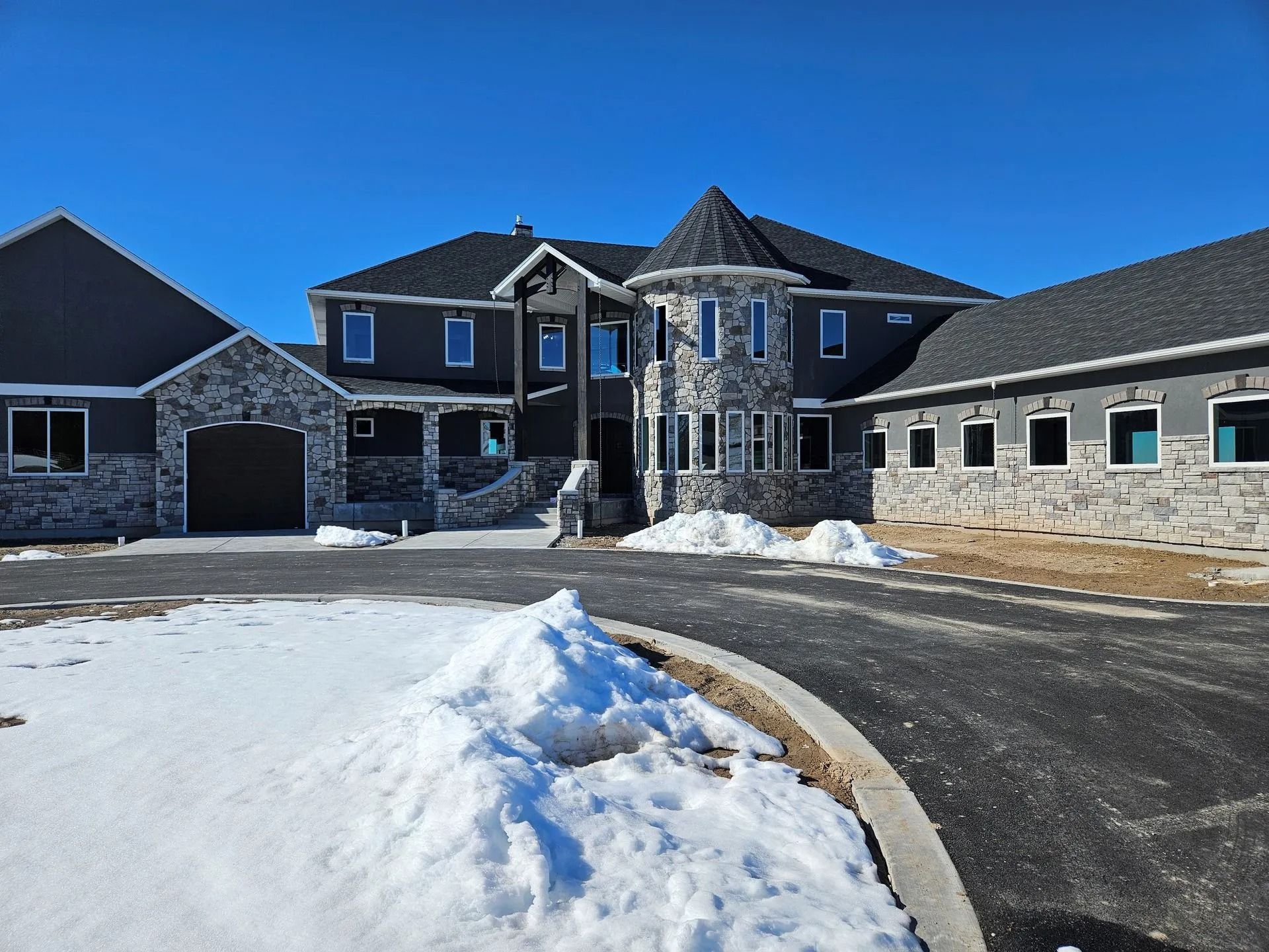 Large two-story house with gray and stone facade, driveway, and patches of snow. Blue sky in the background.