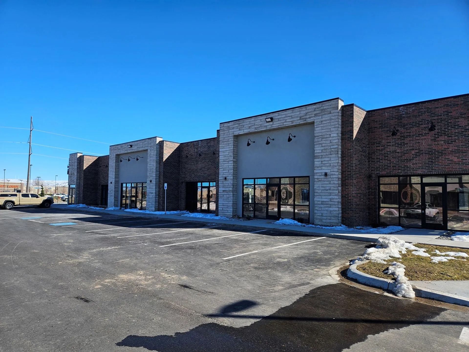 Exterior view of a commercial building with brick and tan stone facade, several storefronts, and a paved parking lot.