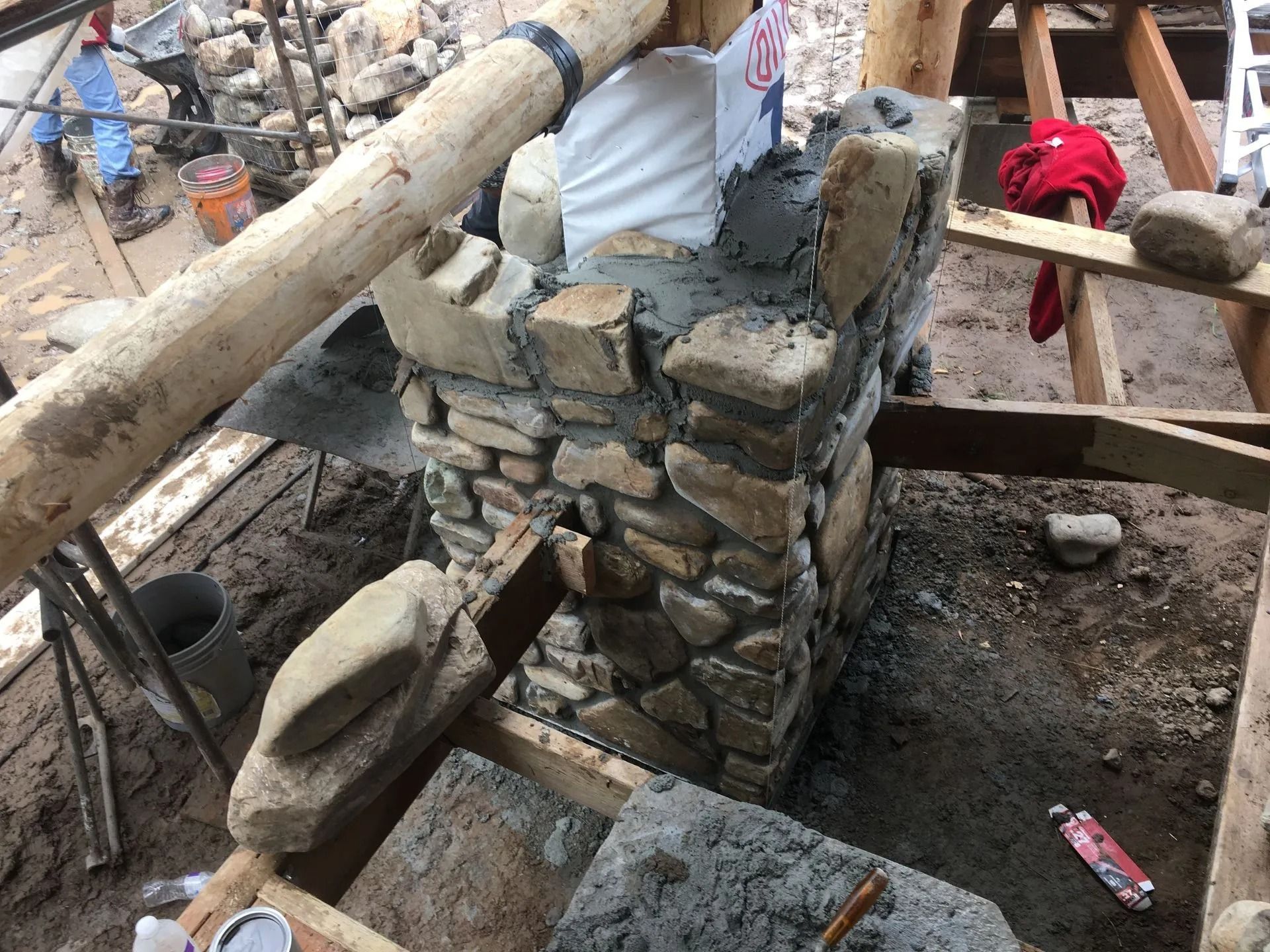 Stone chimney construction in progress; wooden beams and supports surround the structure.