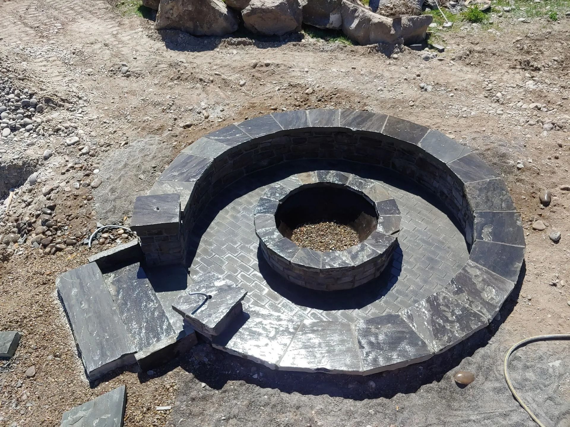 A partially constructed, spiral-shaped stone structure with a central fire pit in an outdoor setting.