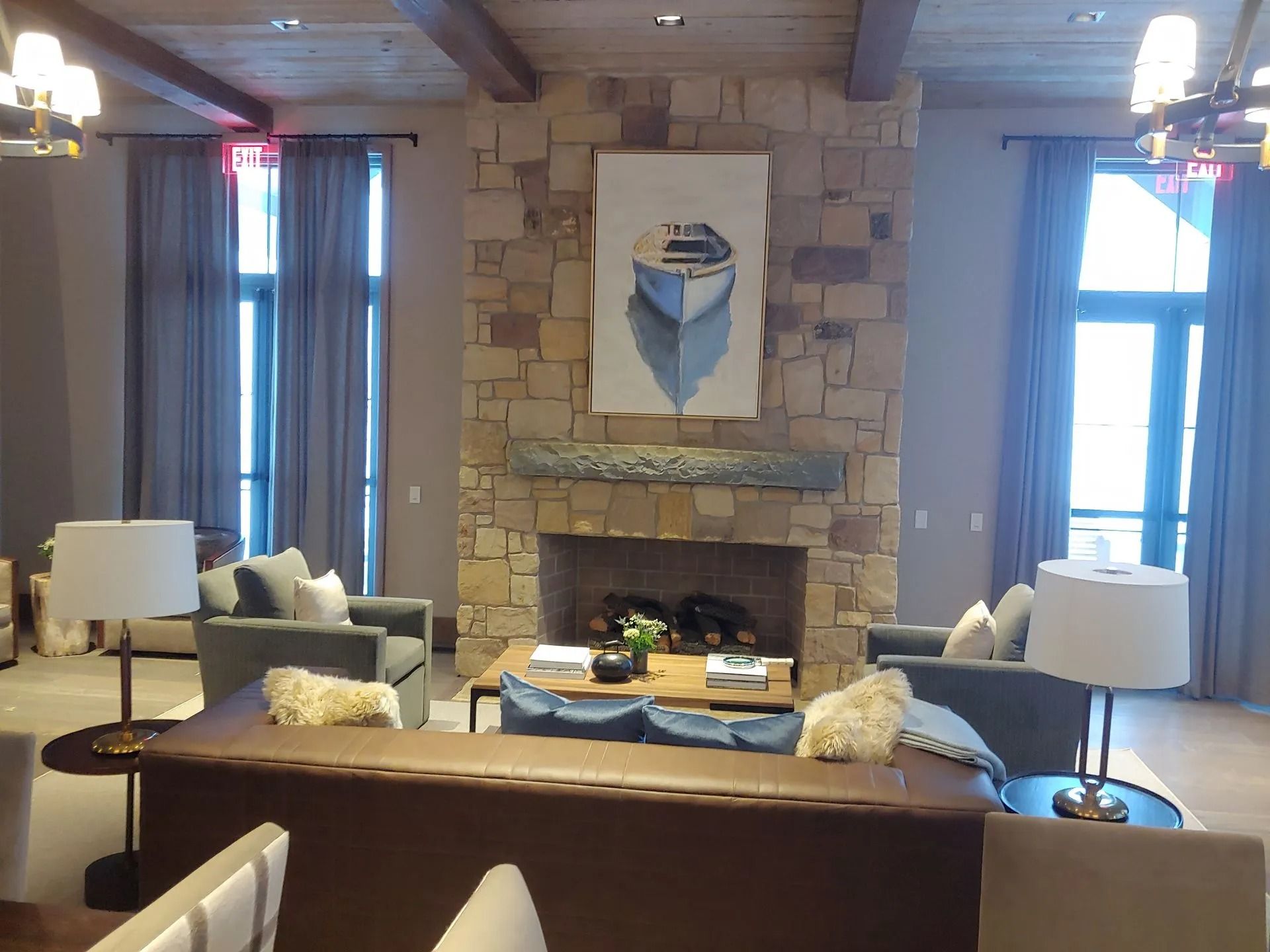 Cozy room with a stone fireplace, boat art, and seating by large windows with blue curtains.