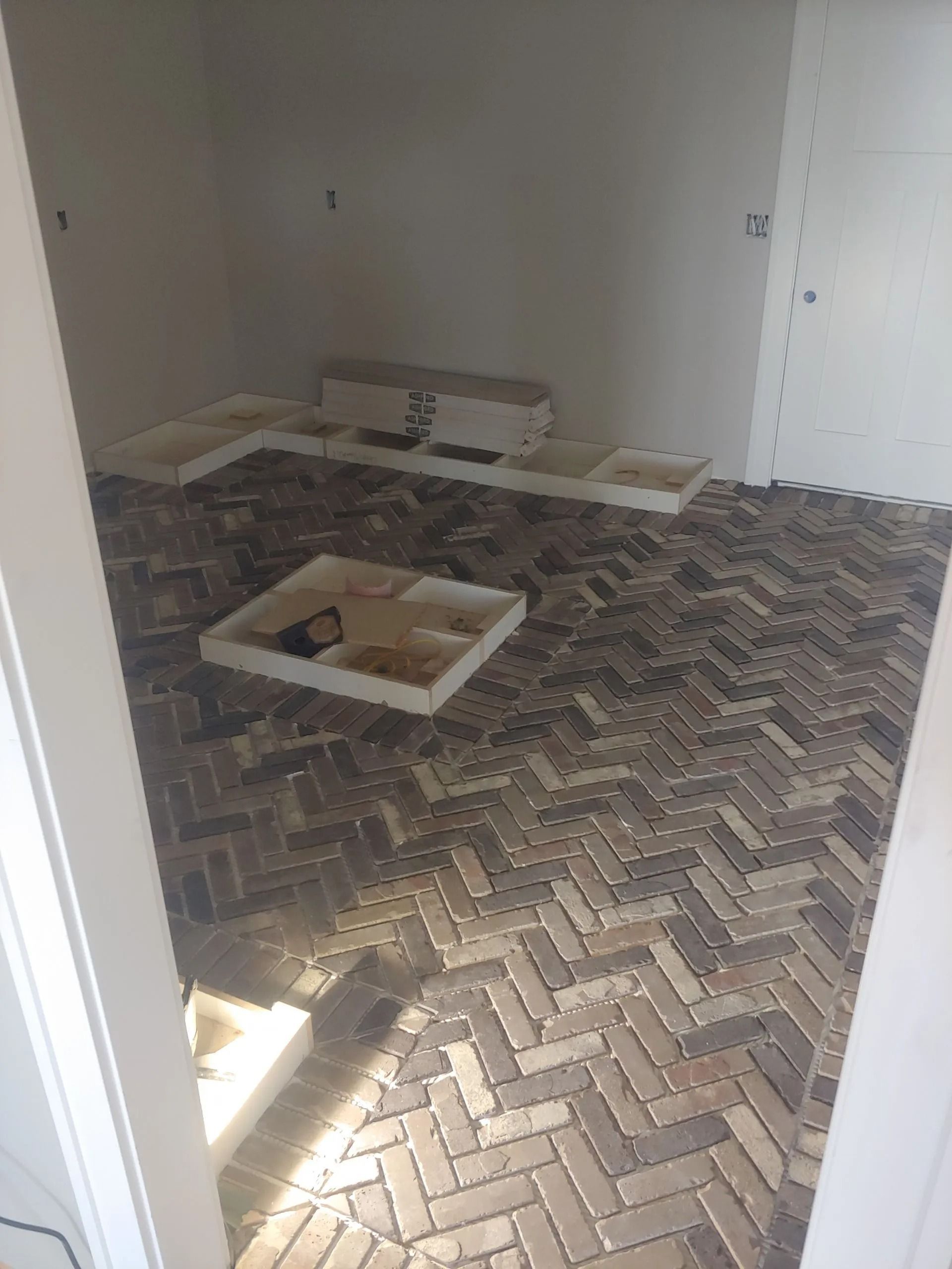 Herringbone brick floor in a room under construction, with white storage trays and a white wall.