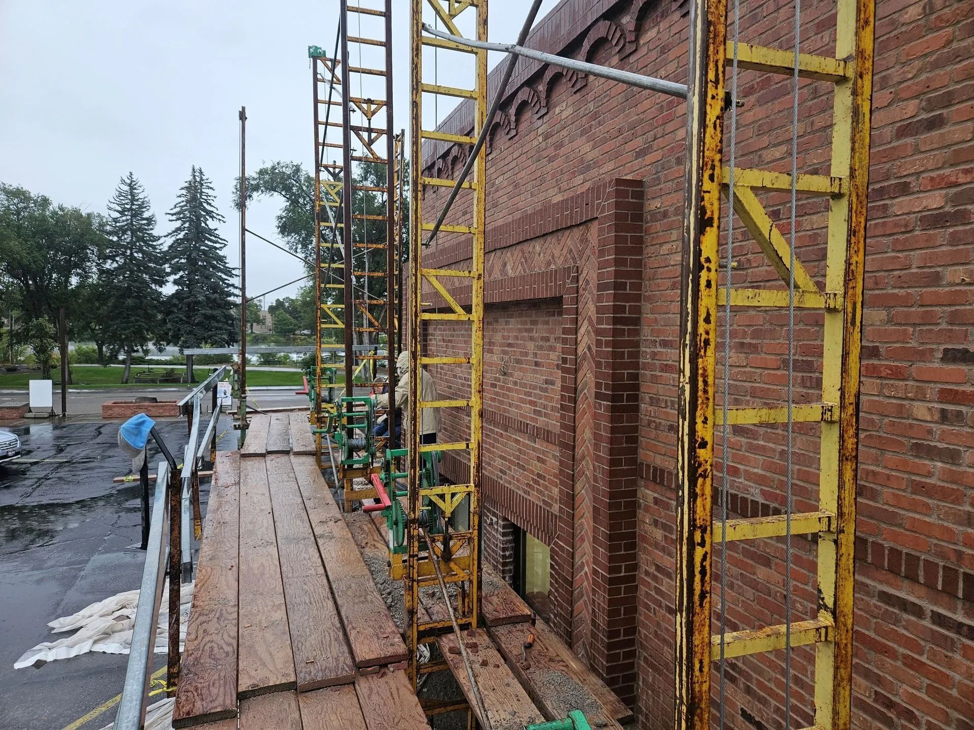 Scaffolding along brick building exterior. Wooden planks, yellow metal supports. Cloudy sky, trees in background.