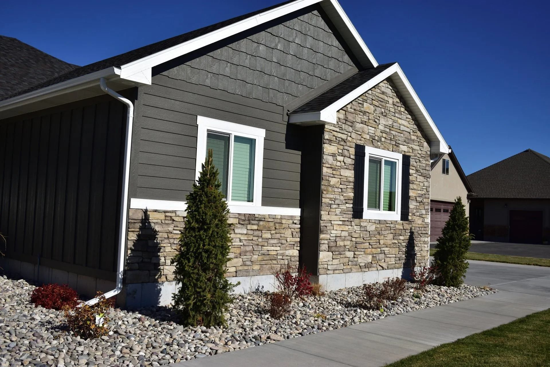 Gray and stone-faced house with white trim, a walkway, and shrubbery on a sunny day.