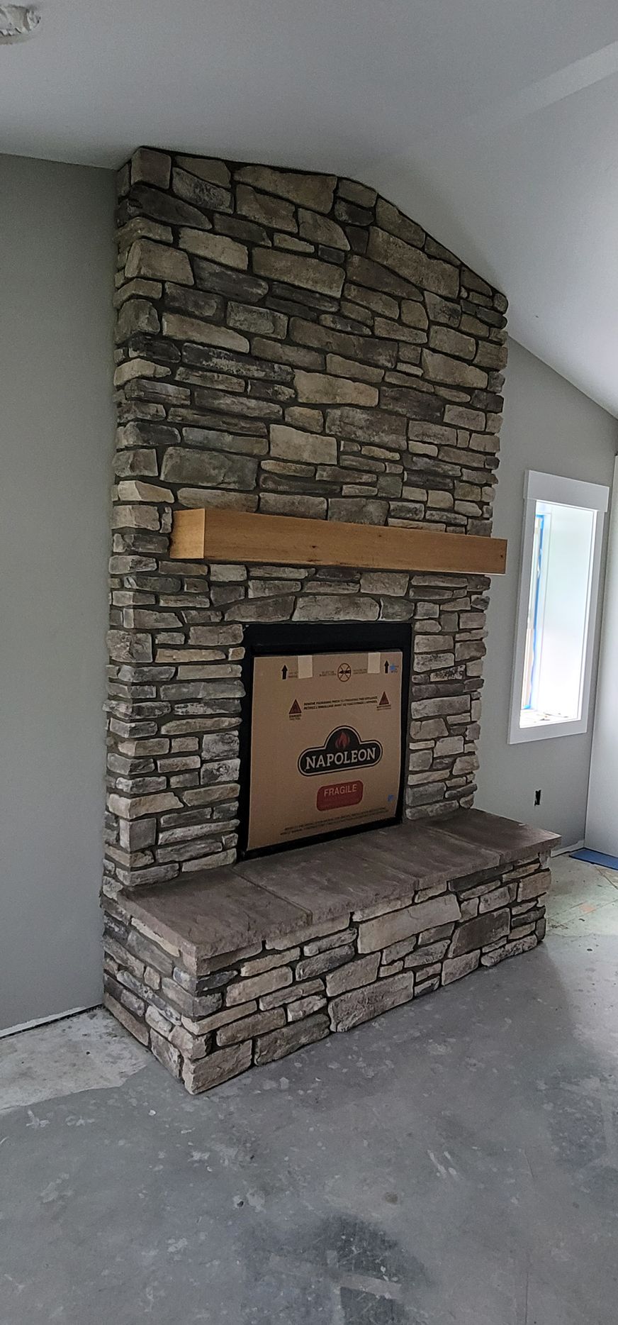 Stone fireplace with wooden mantel, set in a room with gray walls and a window.