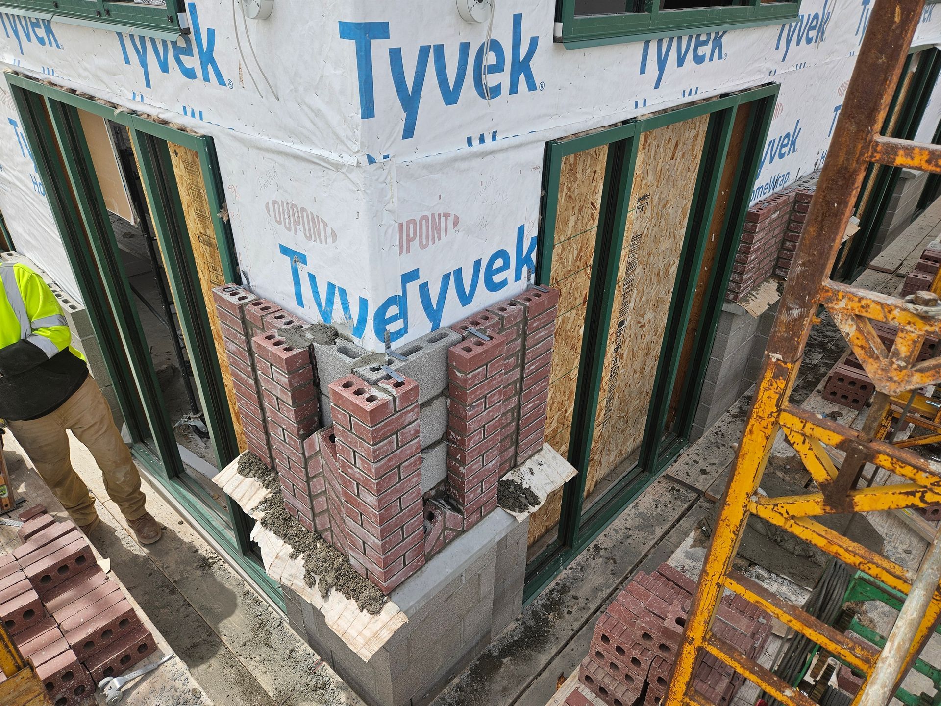 Corner of a building under construction, brickwork in progress. Tyvek wrap, green trim, ladder, and worker.