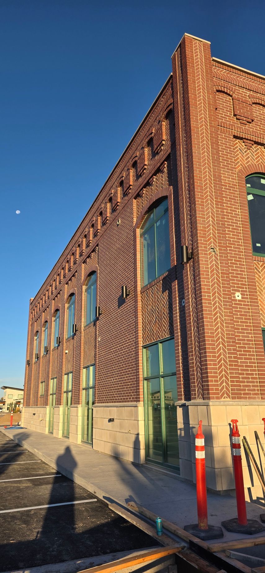 Brick building with arched windows and a blue sky. There are orange and white traffic pylons and a shadow on the concrete sidewalk.