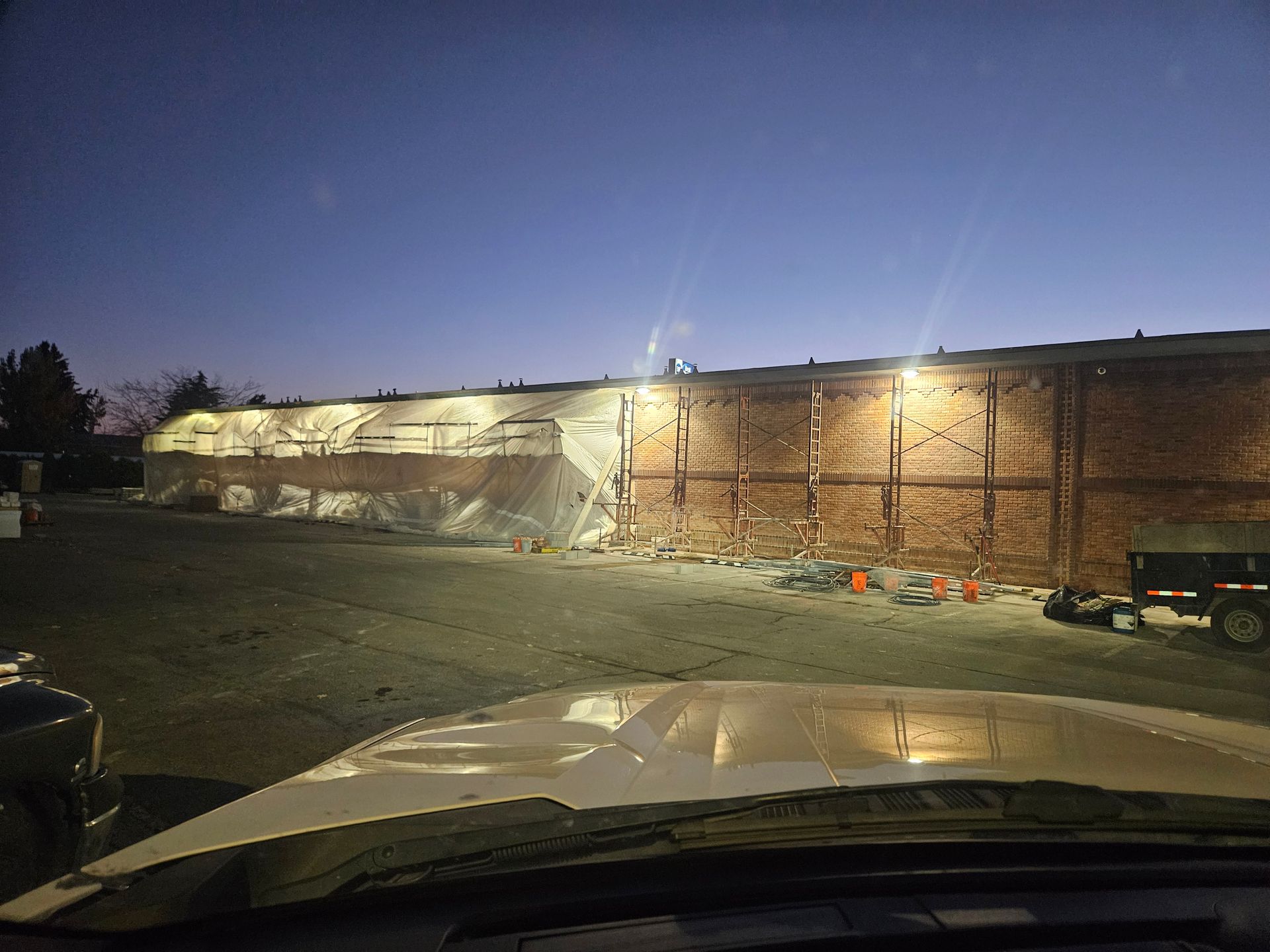 Brick building under construction, illuminated at dusk. Part of wall removed, with safety cones and equipment nearby.