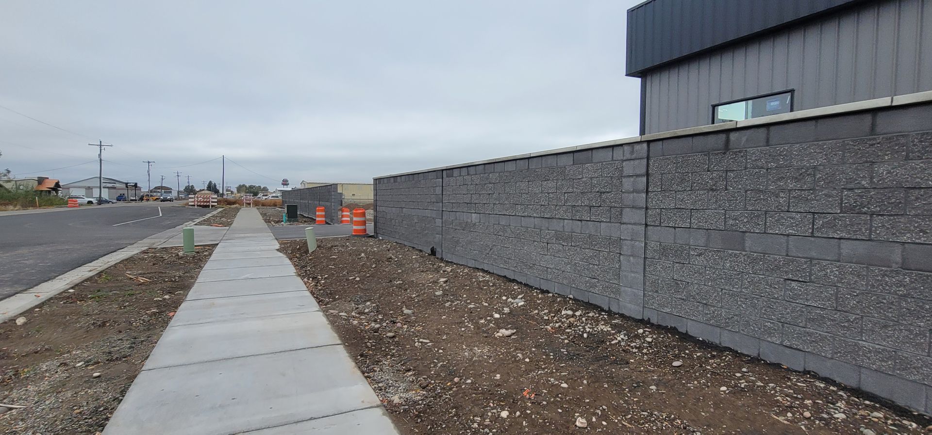 Sidewalk next to a gravel wall and a paved road with parked cars and orange cones under a cloudy sky.
