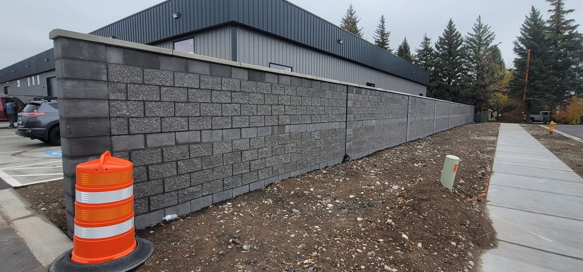 A tall, gray brick wall borders a parking lot and sidewalk, with an orange traffic cone in the foreground.