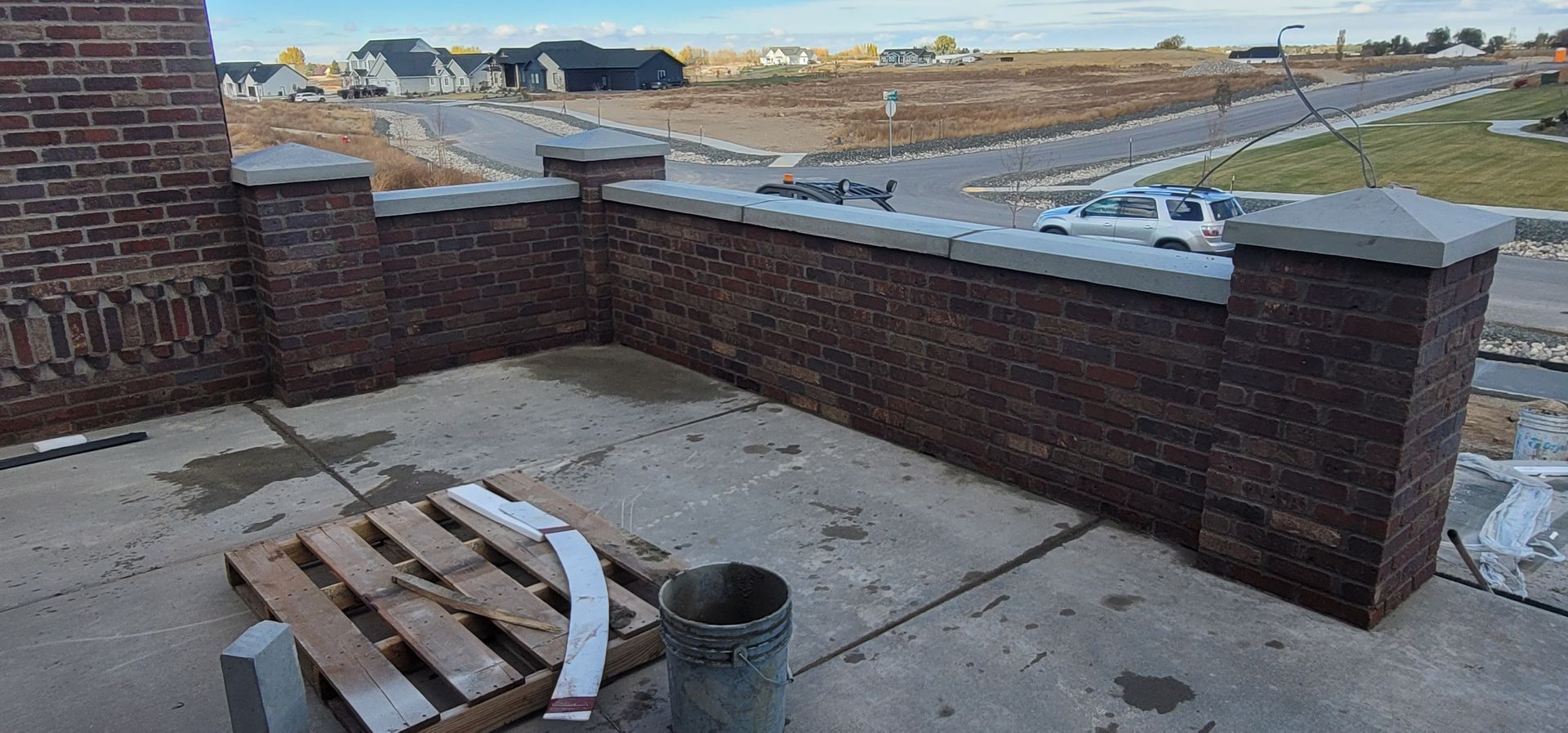 Brick wall with concrete cap, posts, and nearby construction materials. New neighborhood in background.