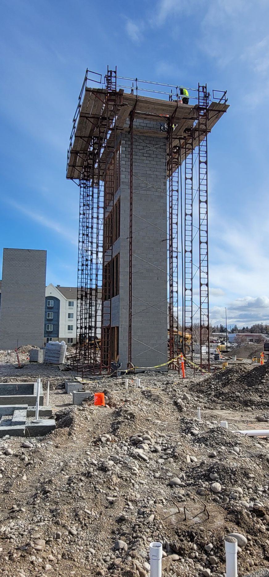 Construction site with tall brick structure surrounded by scaffolding, workers on top, blue sky background.