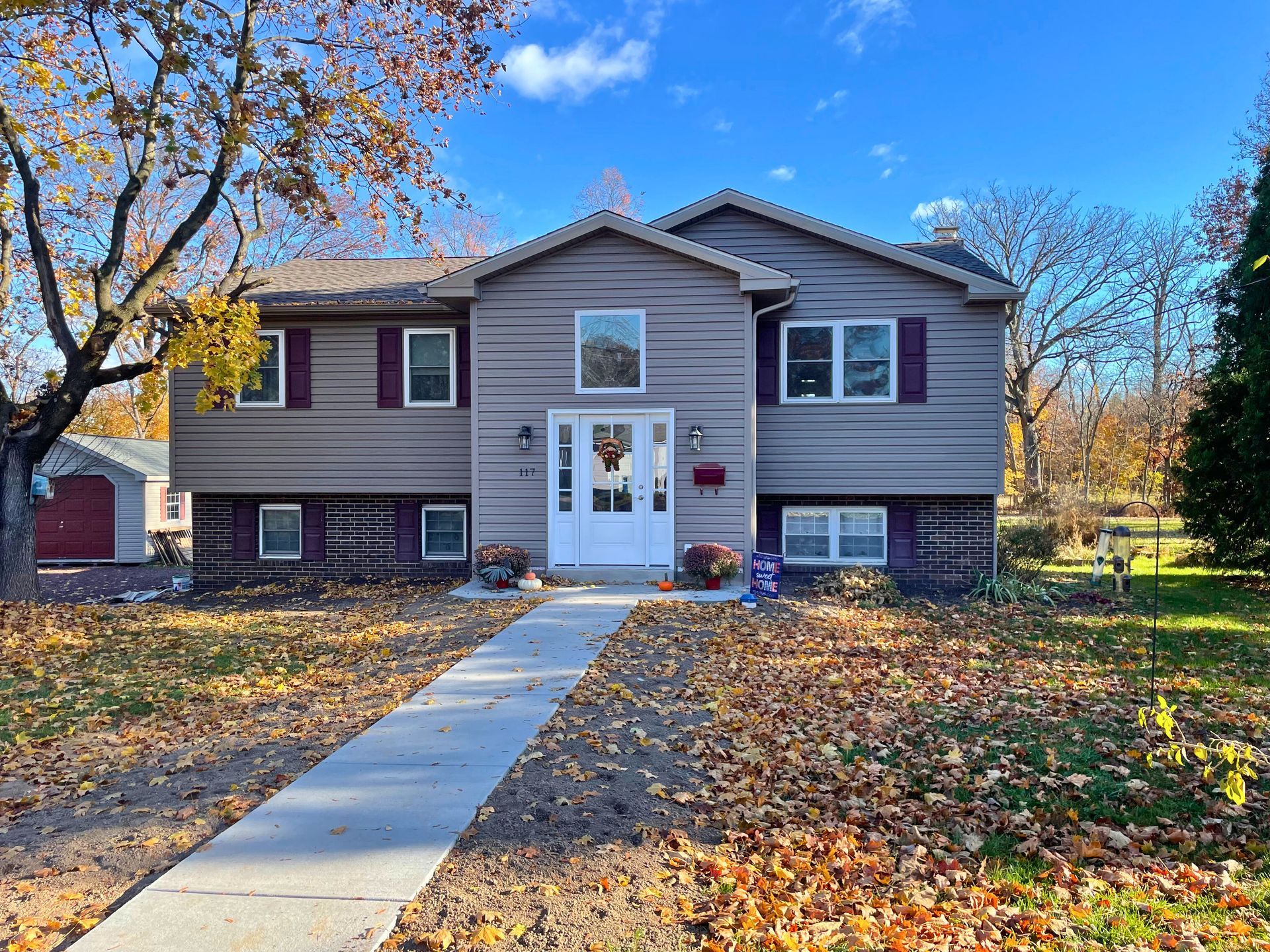 Two-story house with brown siding, a white door, and burgundy shutters on a sunny autumn day.