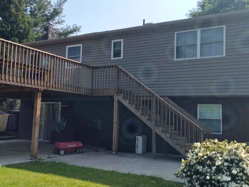 Two-story house with wooden deck and stairs, gray siding. Lower level is a patio. Green lawn in the foreground.