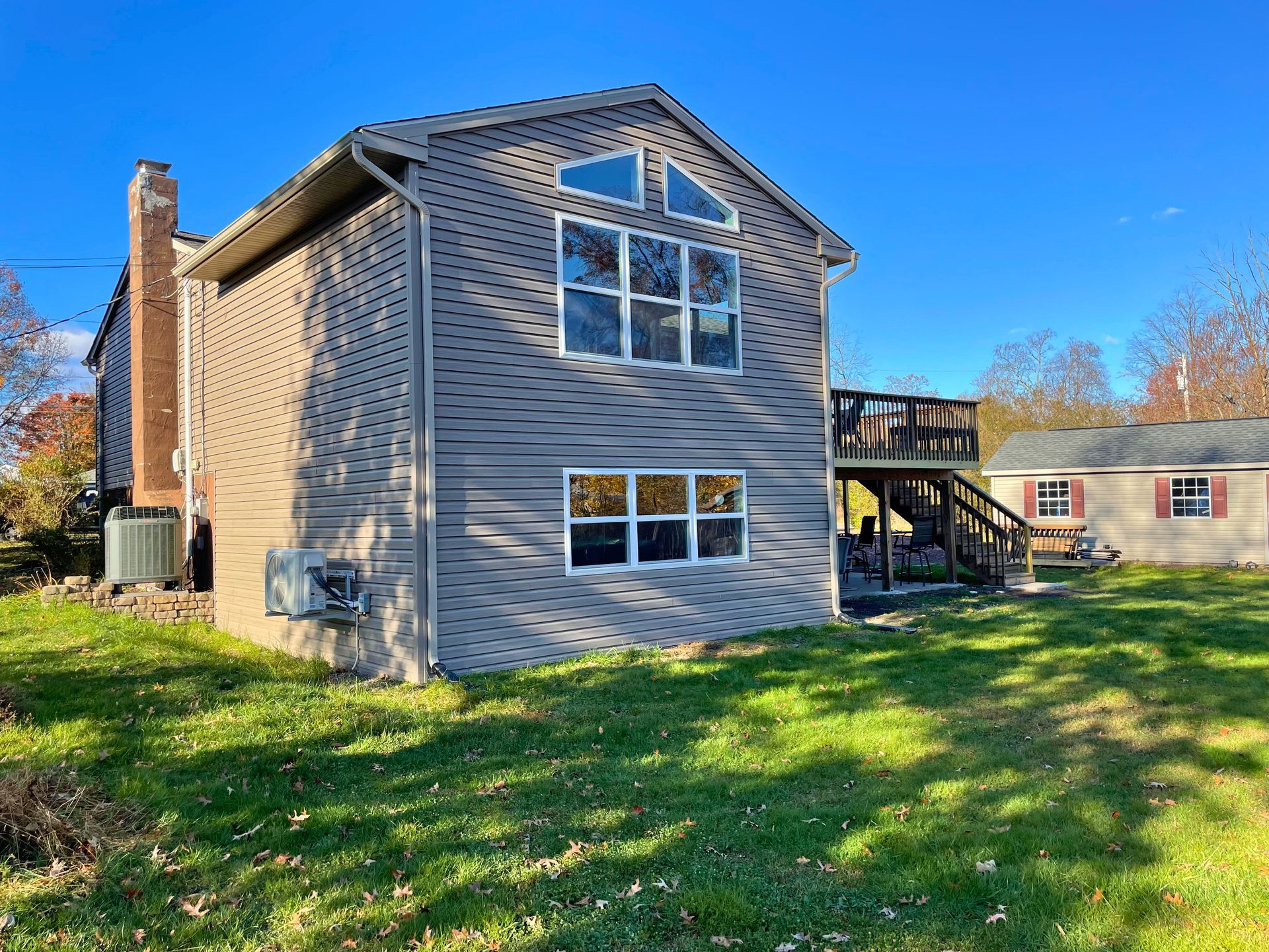 Side view of a two-story house with gray siding, a deck, and a detached shed in a grassy yard on a sunny day.