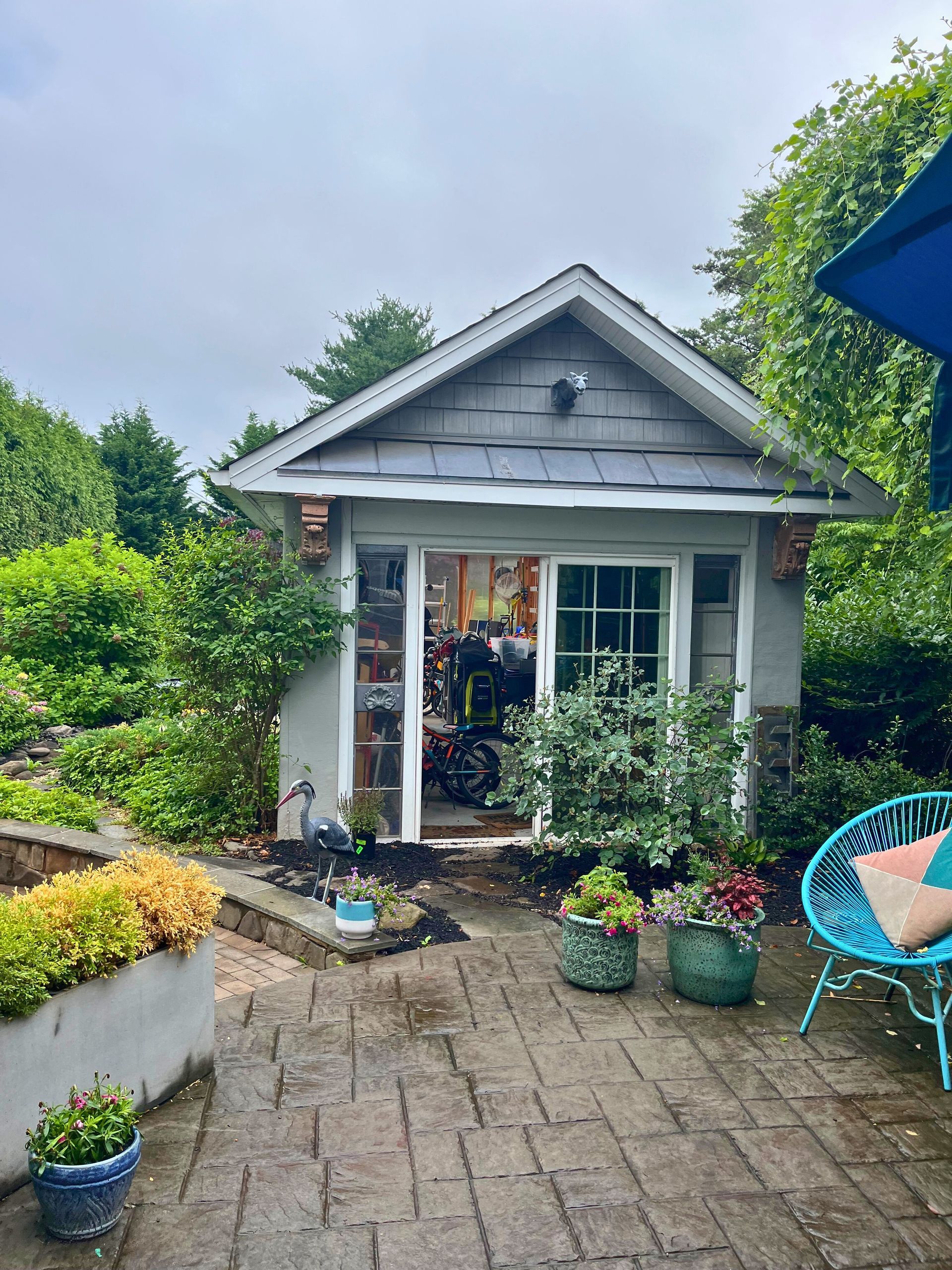 Small gray garden shed with glass doors and plants on a brick patio.