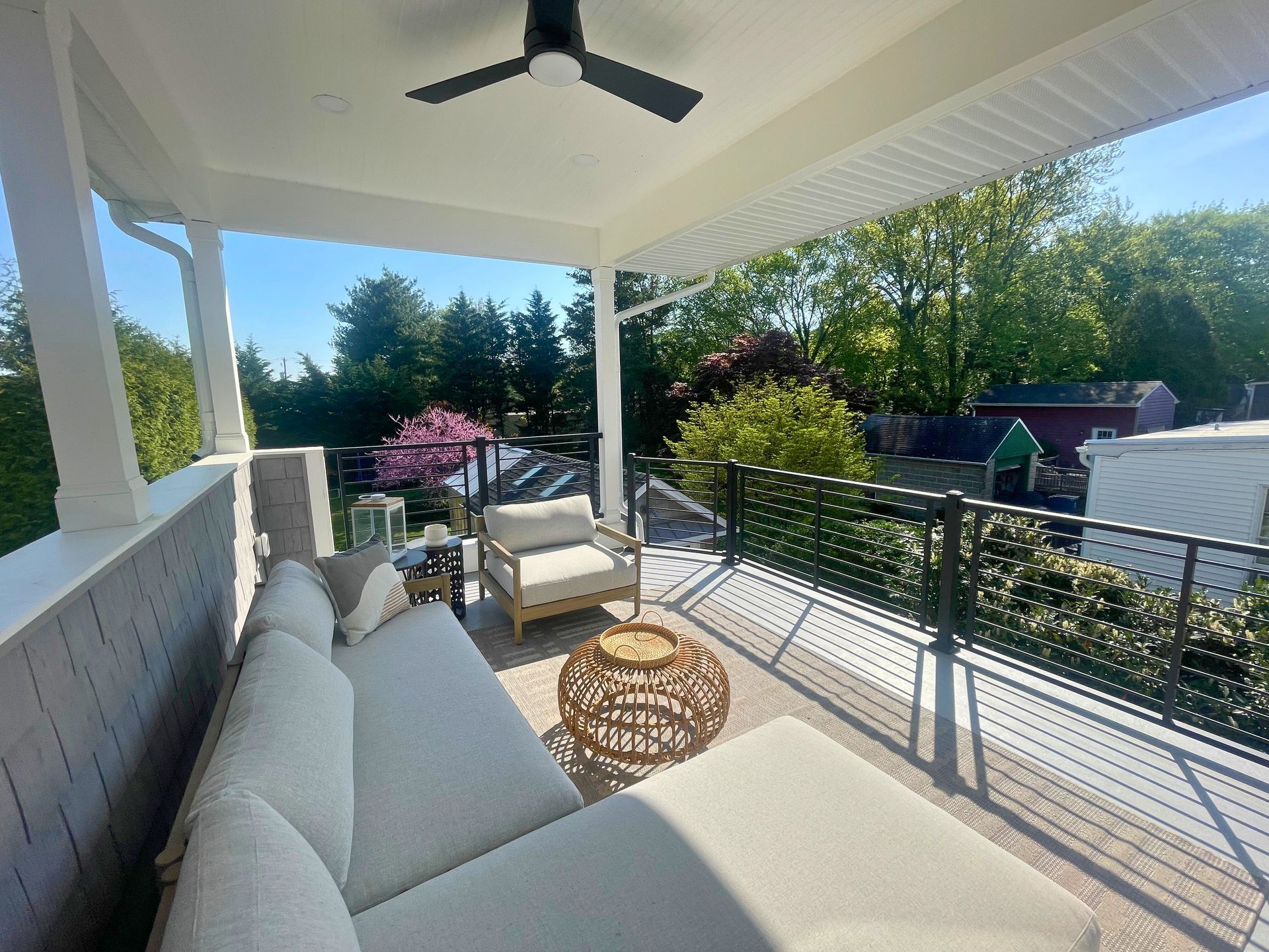 A covered outdoor patio with white sofas, wicker coffee table, and views of trees under a sunny sky.