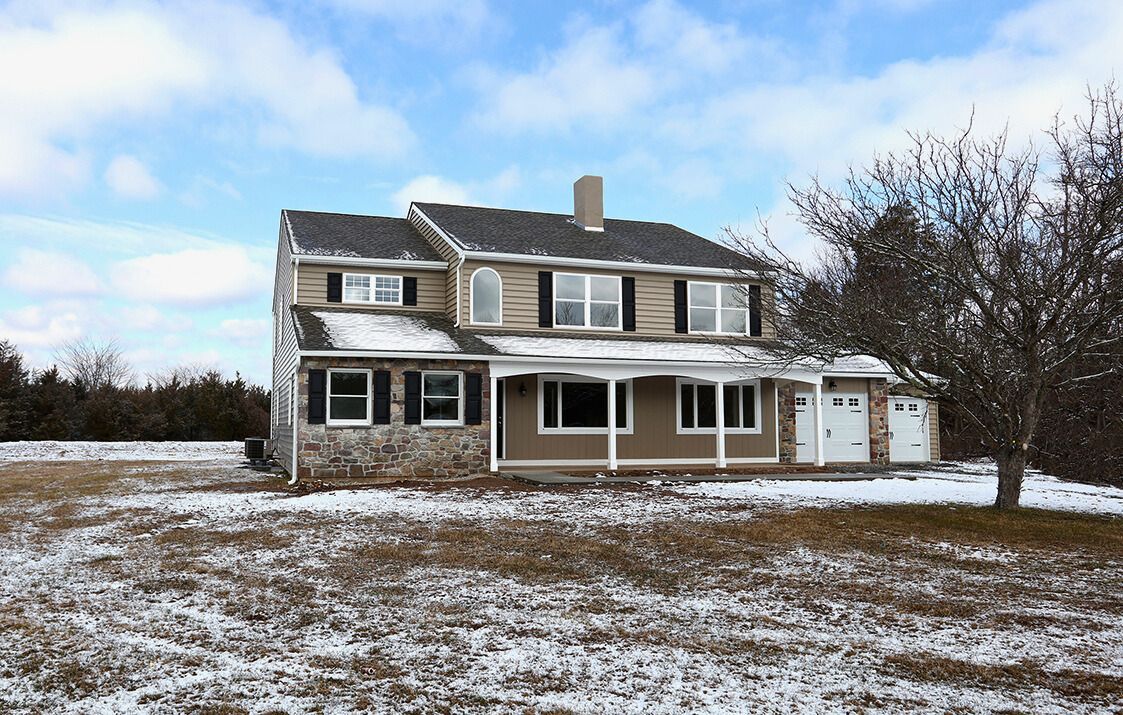 Two-story house with snow-covered roof, brown siding, and stone base in a field with a snowy ground.