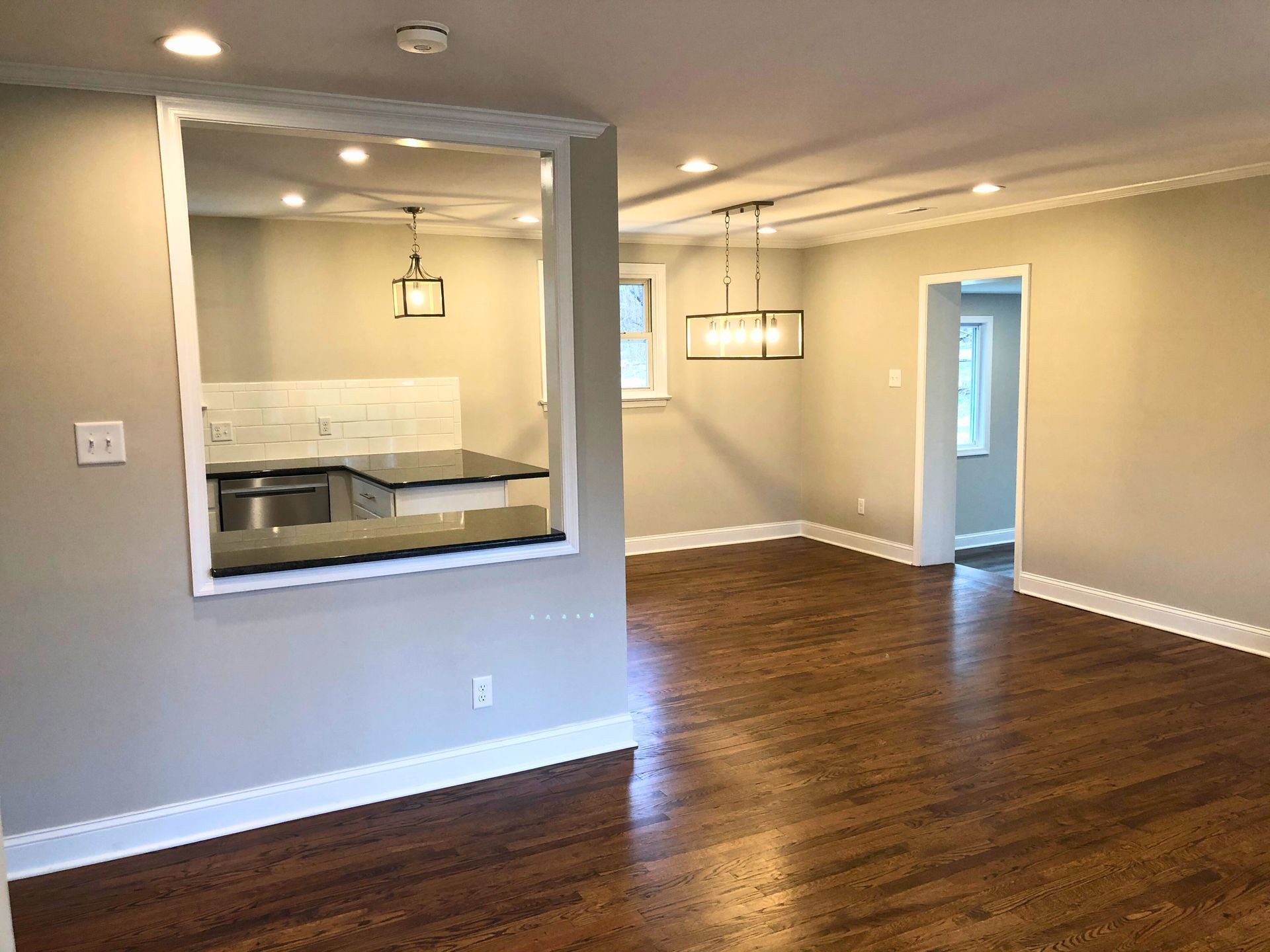 Interior view of a remodeled living room and kitchen with hardwood floors, light gray walls, and recessed lighting.