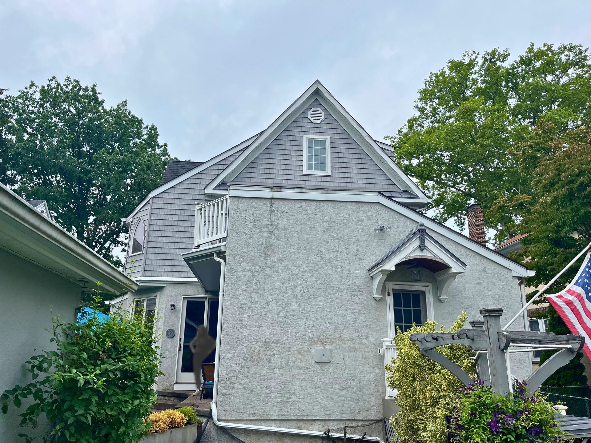 Two-story stucco house with gray roof, small balcony, American flag, and surrounding trees under an overcast sky.