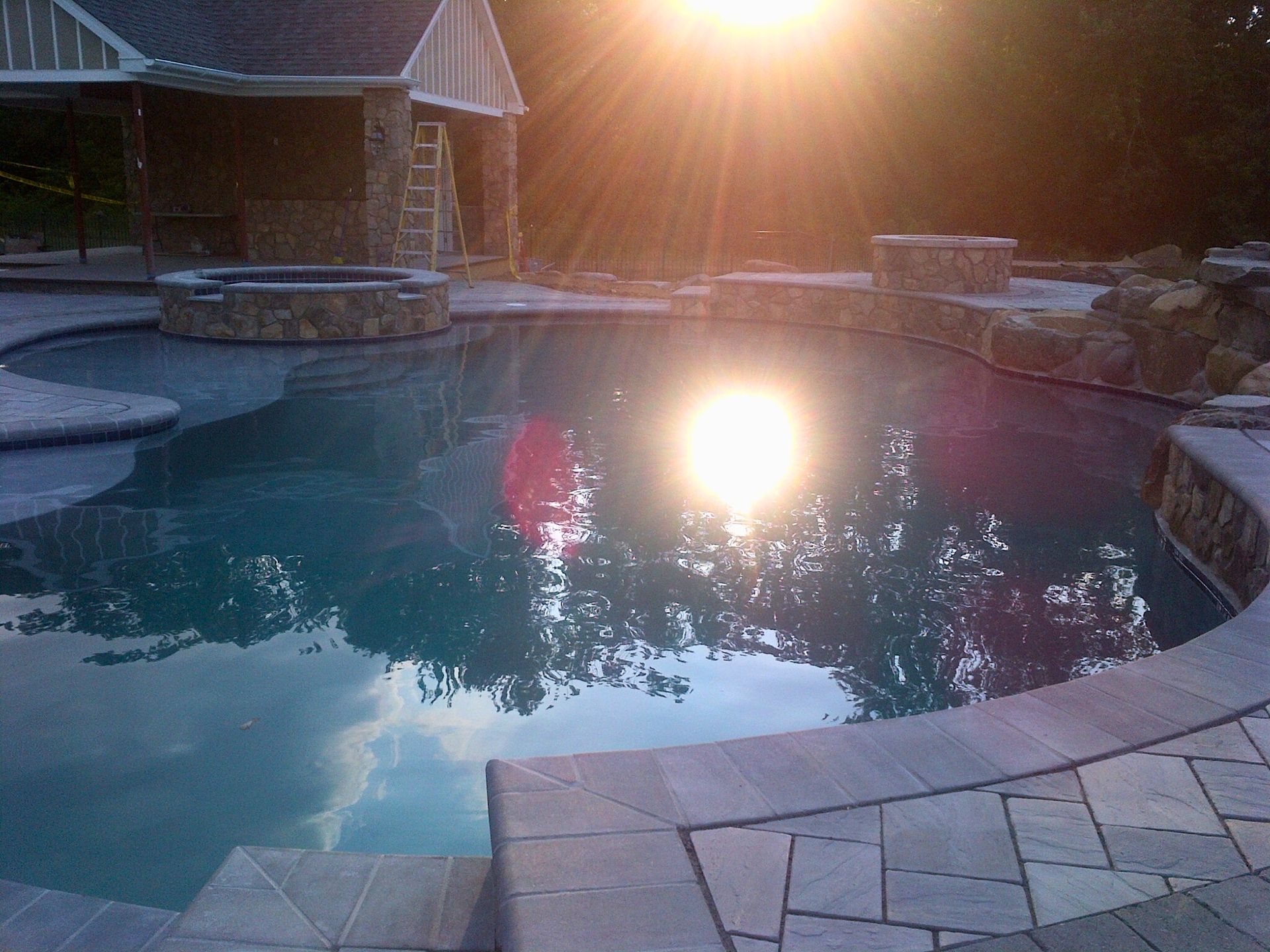Swimming pool reflecting the bright sun, with a stone-covered structure and patio surrounding the water.