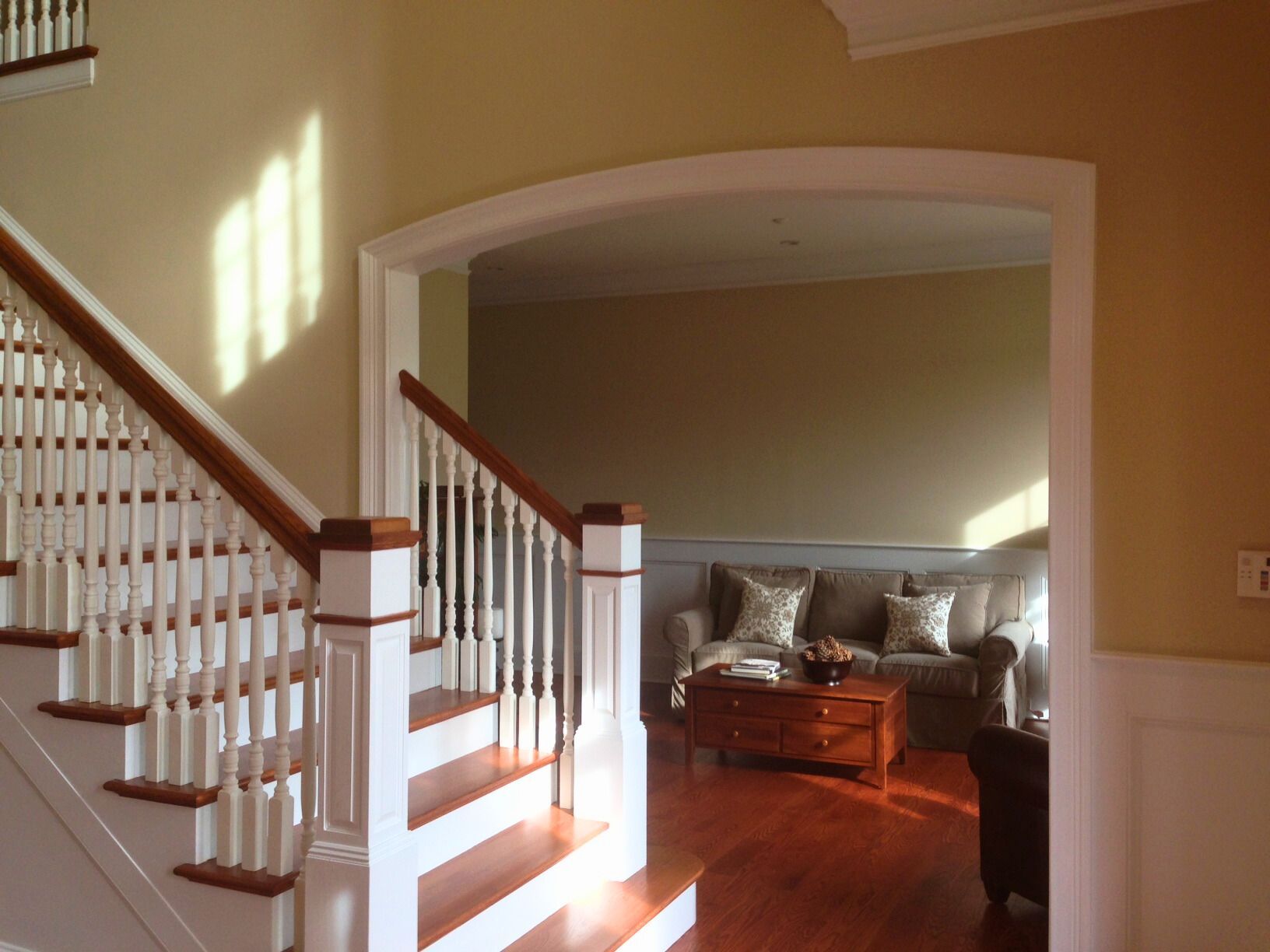 A staircase leads to an archway into a living room with a couch and coffee table; warm sunlight.