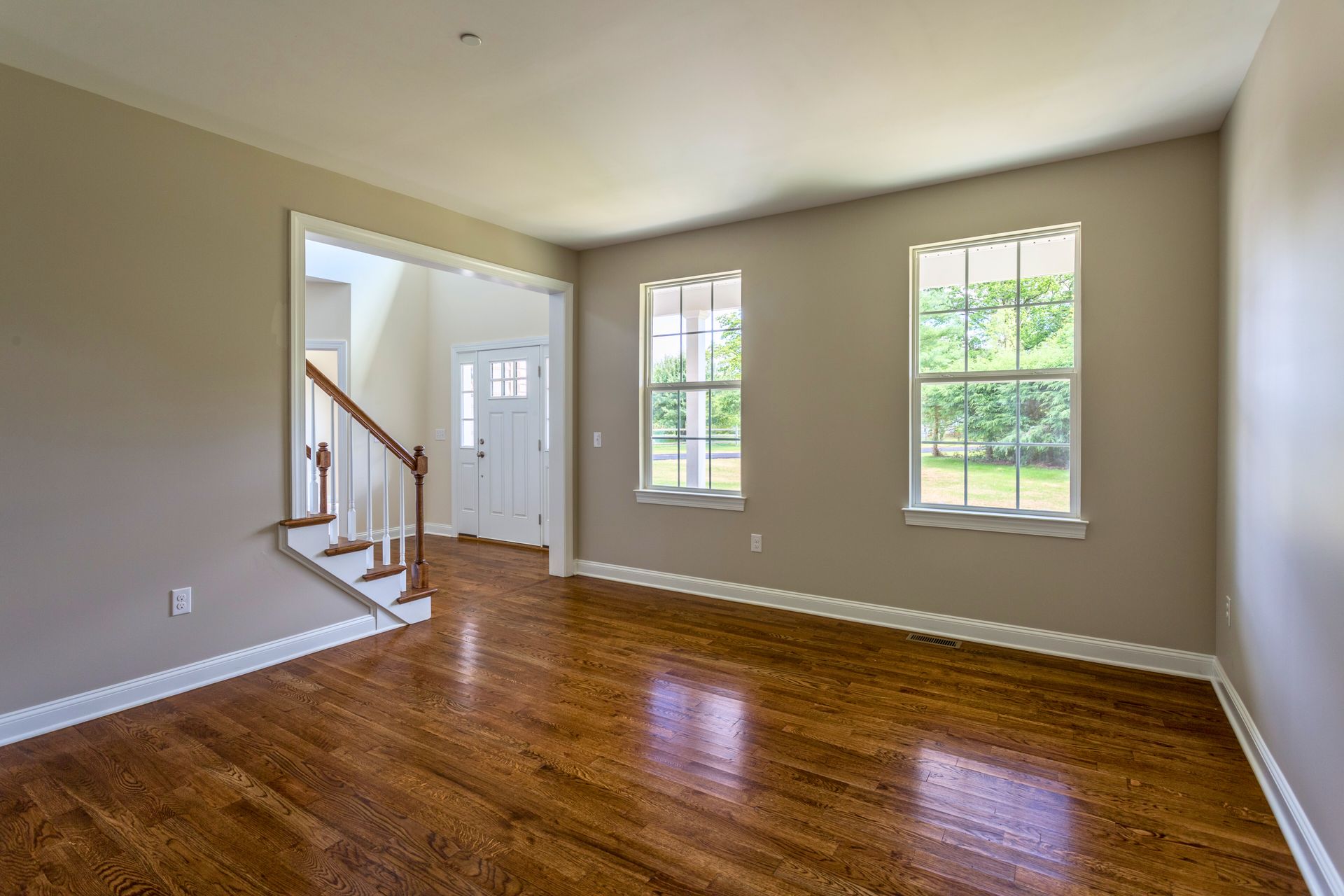 Empty room with hardwood floor, two windows, stairs and door. Beige walls.