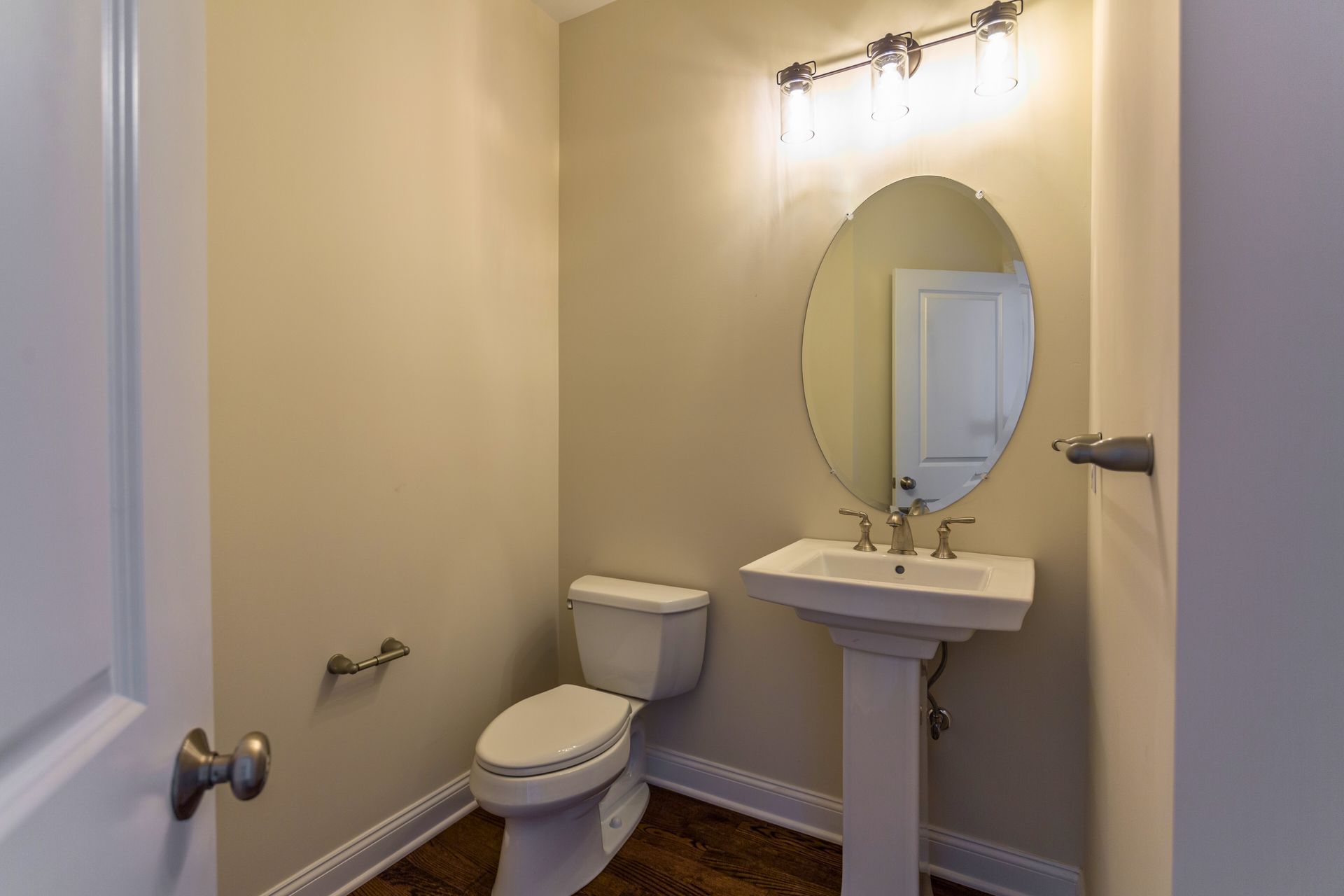 Powder room with toilet, pedestal sink, oval mirror, and light fixture; neutral walls, wood floor.