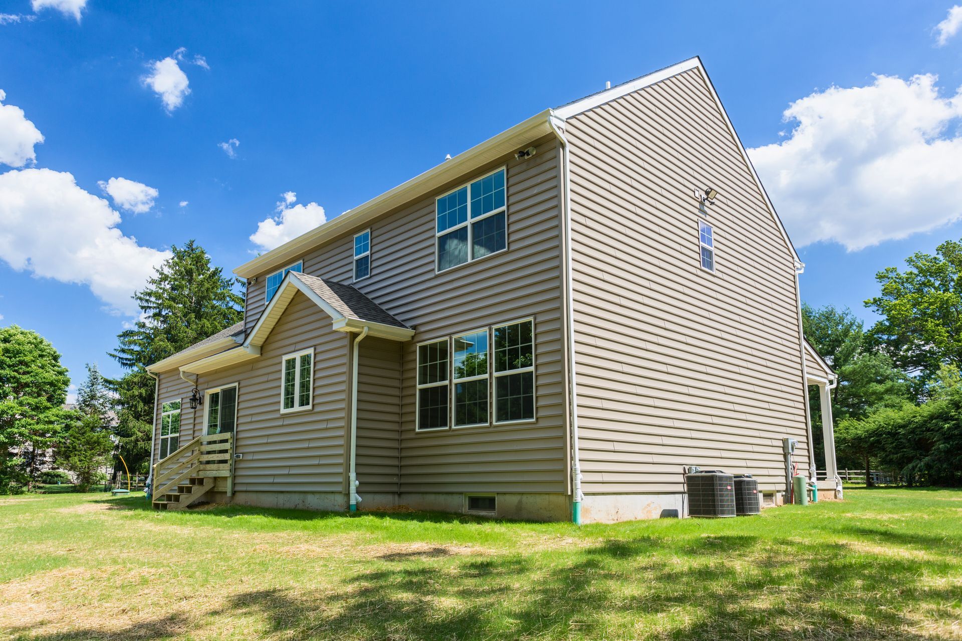 Two-story house with beige siding, windows, and green lawn under a blue sky with clouds.