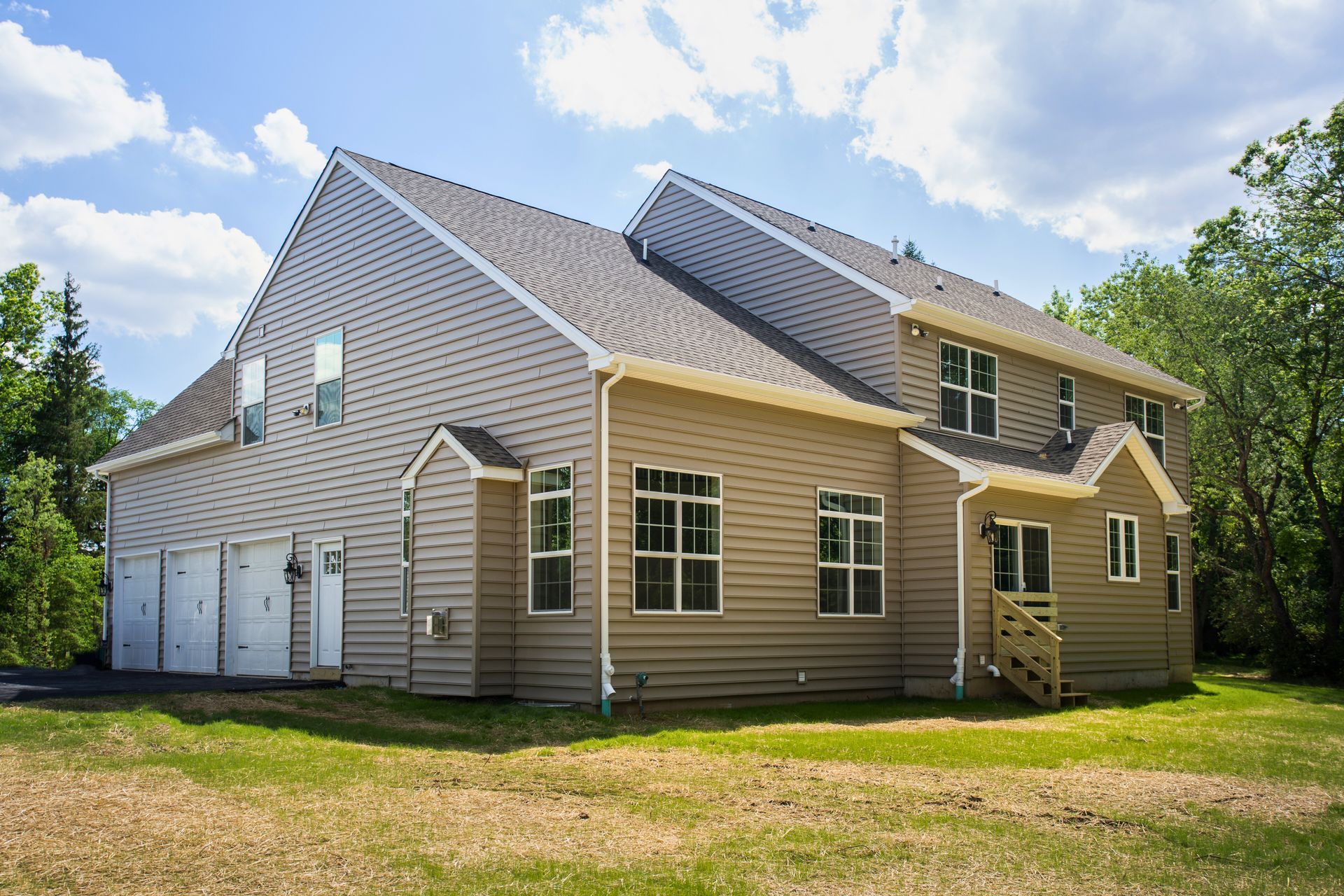 Two-story house with attached garage, tan siding, brown roof, set on green grass under a partly cloudy sky.