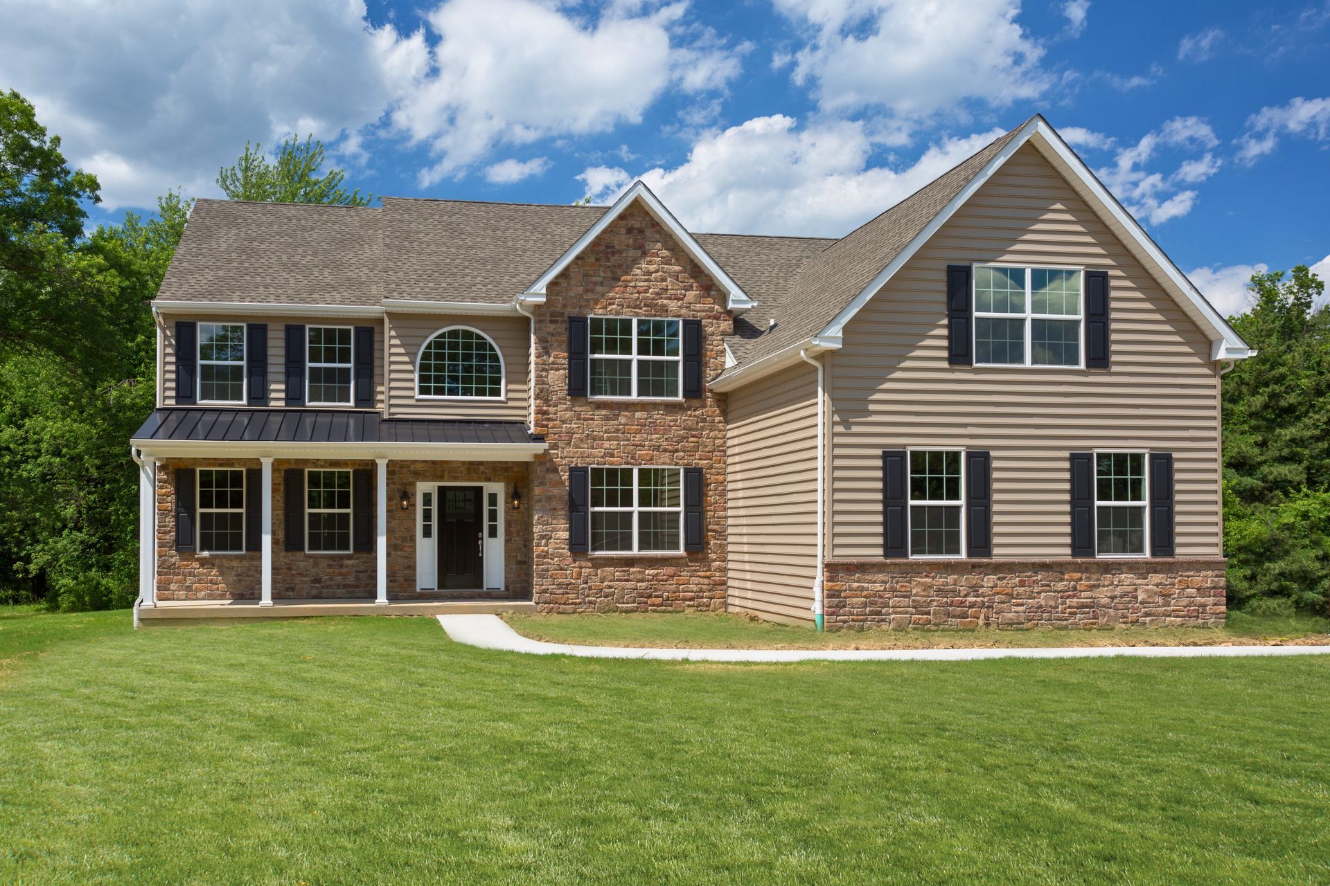 Two-story house with tan siding, brick, and black shutters; green lawn and blue sky.