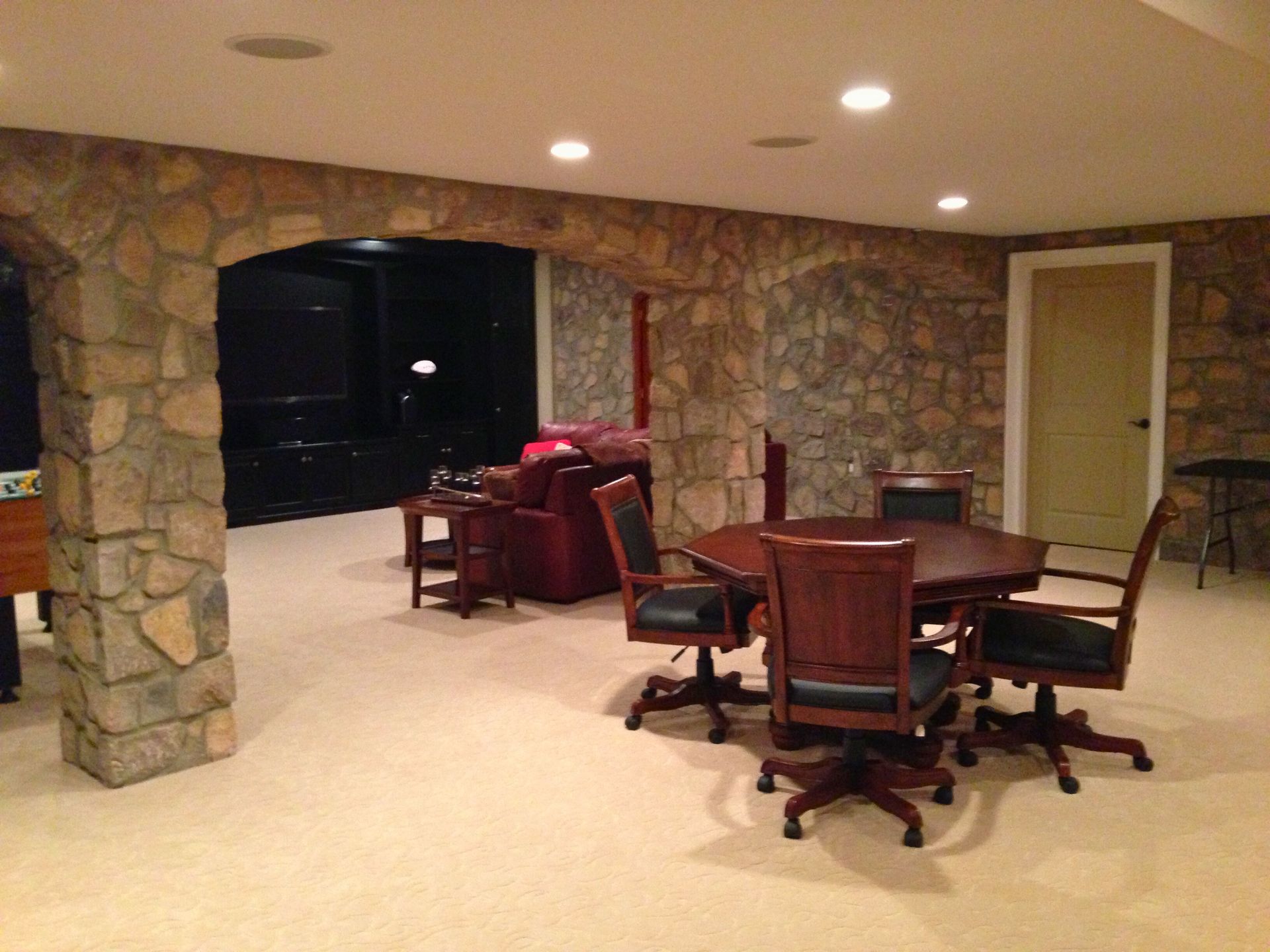 Basement lounge area: stone walls, carpet, round table with chairs, archway to a dark space.