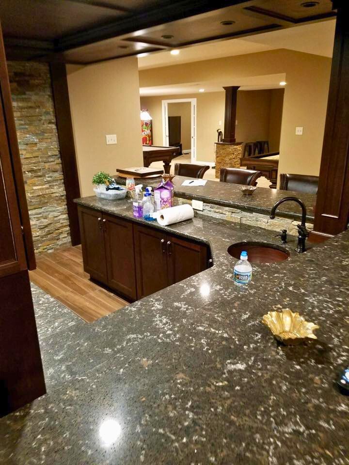 Interior view of a bar with dark cabinets, countertops, and a stone wall. Another room is visible in the background.