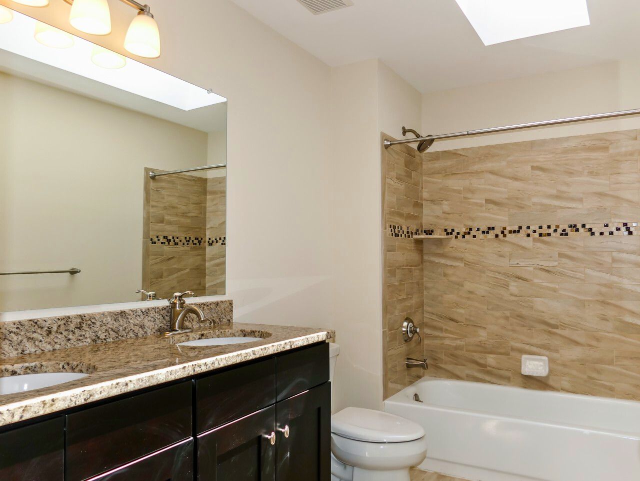 Bathroom with double sink vanity, tub/shower combo, tan tile, and skylight.
