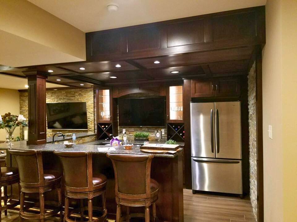Basement bar with dark wood cabinets, stone accents, stainless steel refrigerator, and bar stools.