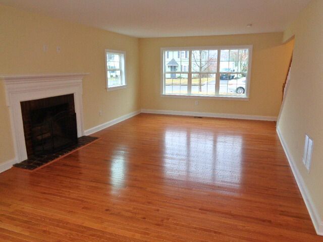 Empty living room with hardwood floors, fireplace, large window, and neutral walls.