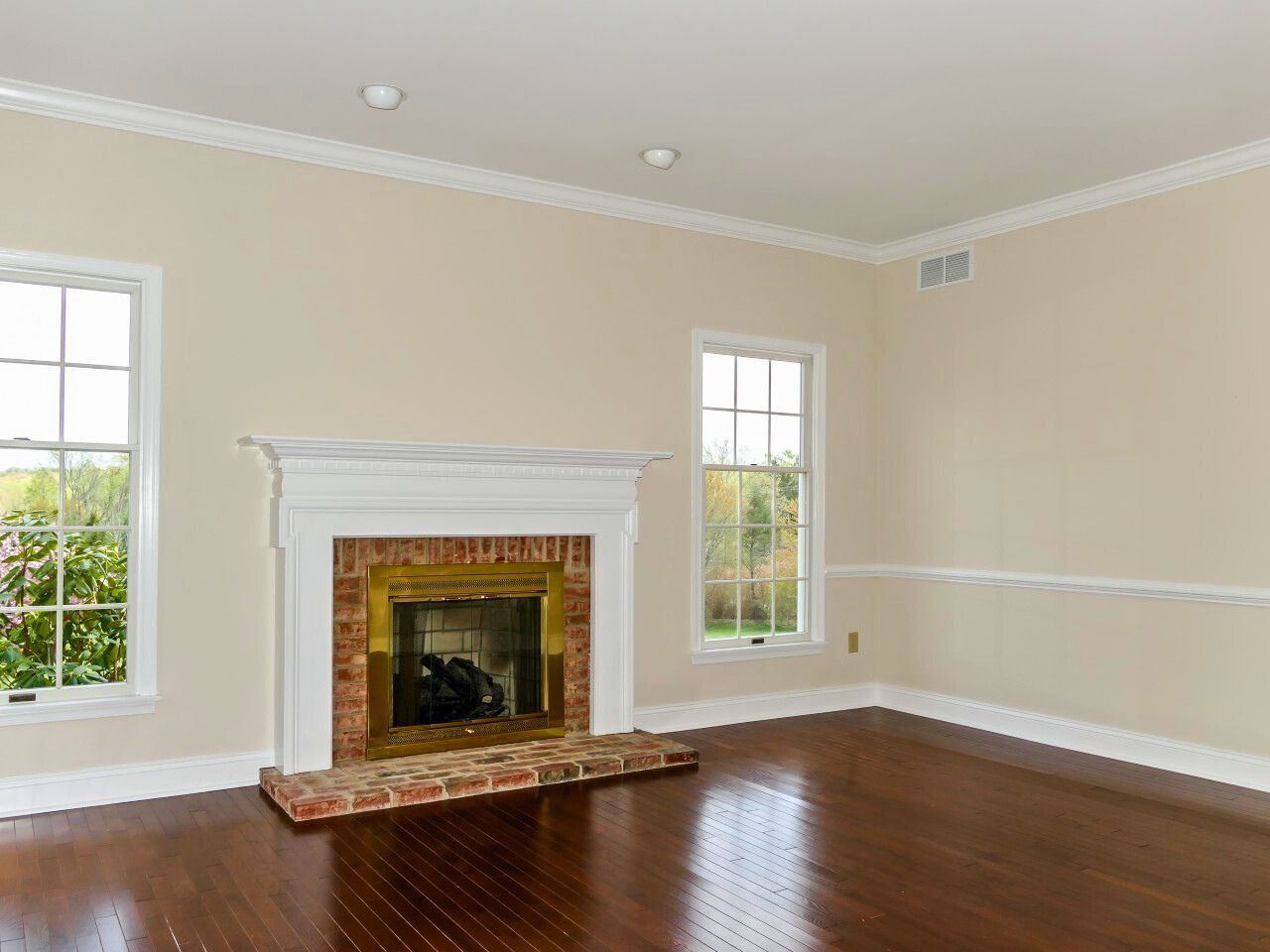 Empty room with fireplace, windows, and wood floors. Beige walls and white trim.
