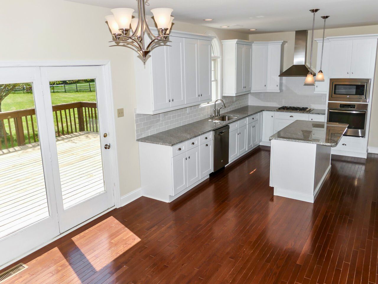 Bright, white kitchen with wooden floors, stainless steel appliances, and doors opening to a deck overlooking a green yard.