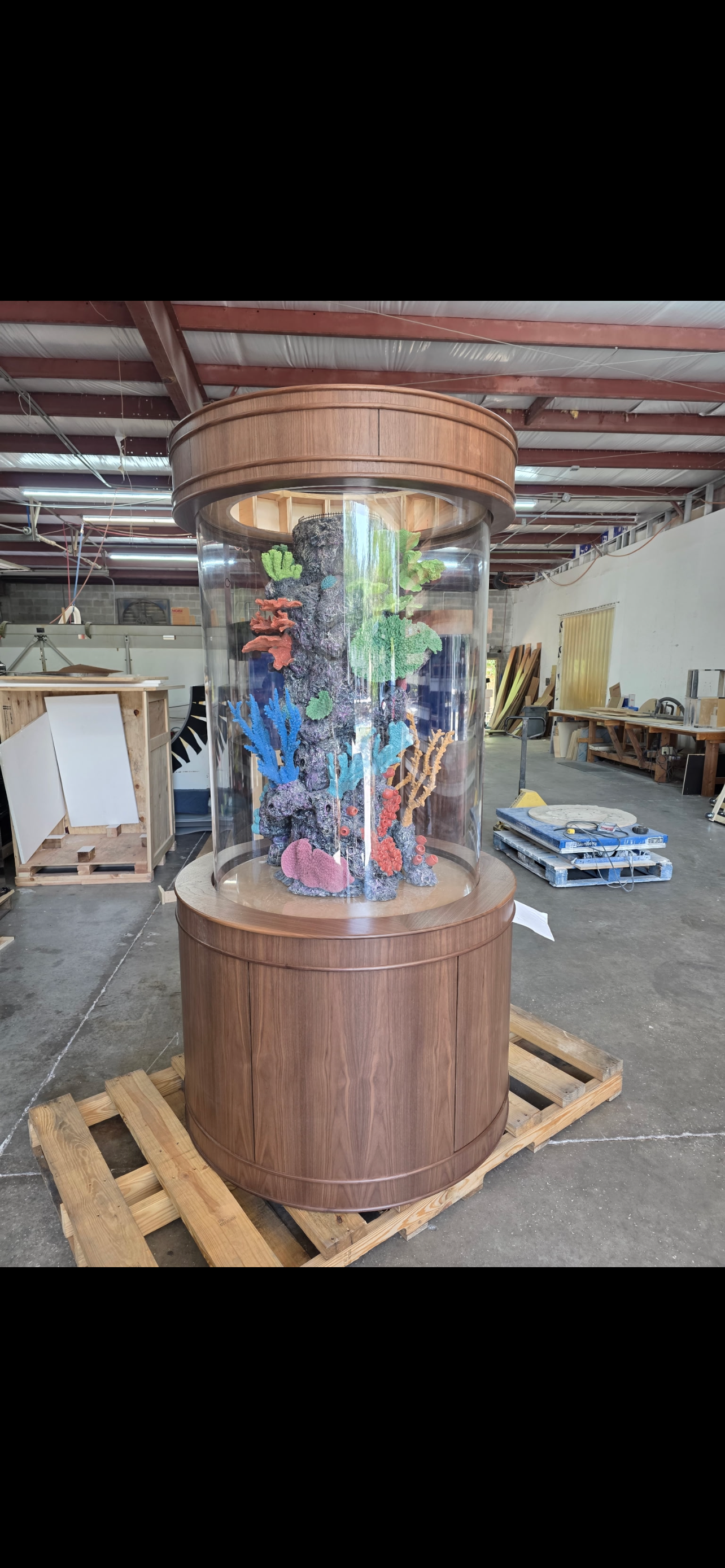A cylindrical aquarium with a decorative coral centerpiece, set inside a wooden base and canopy in a warehouse.