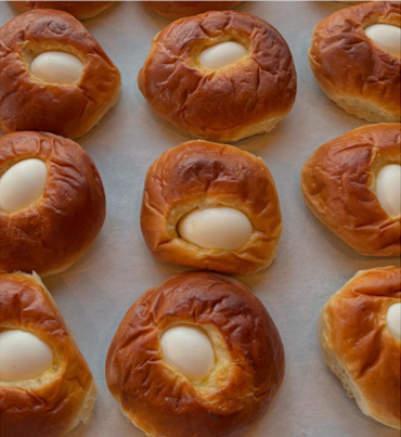 A tray of golden-brown, round baked buns, each featuring a centered, smooth, white hard-boiled egg.