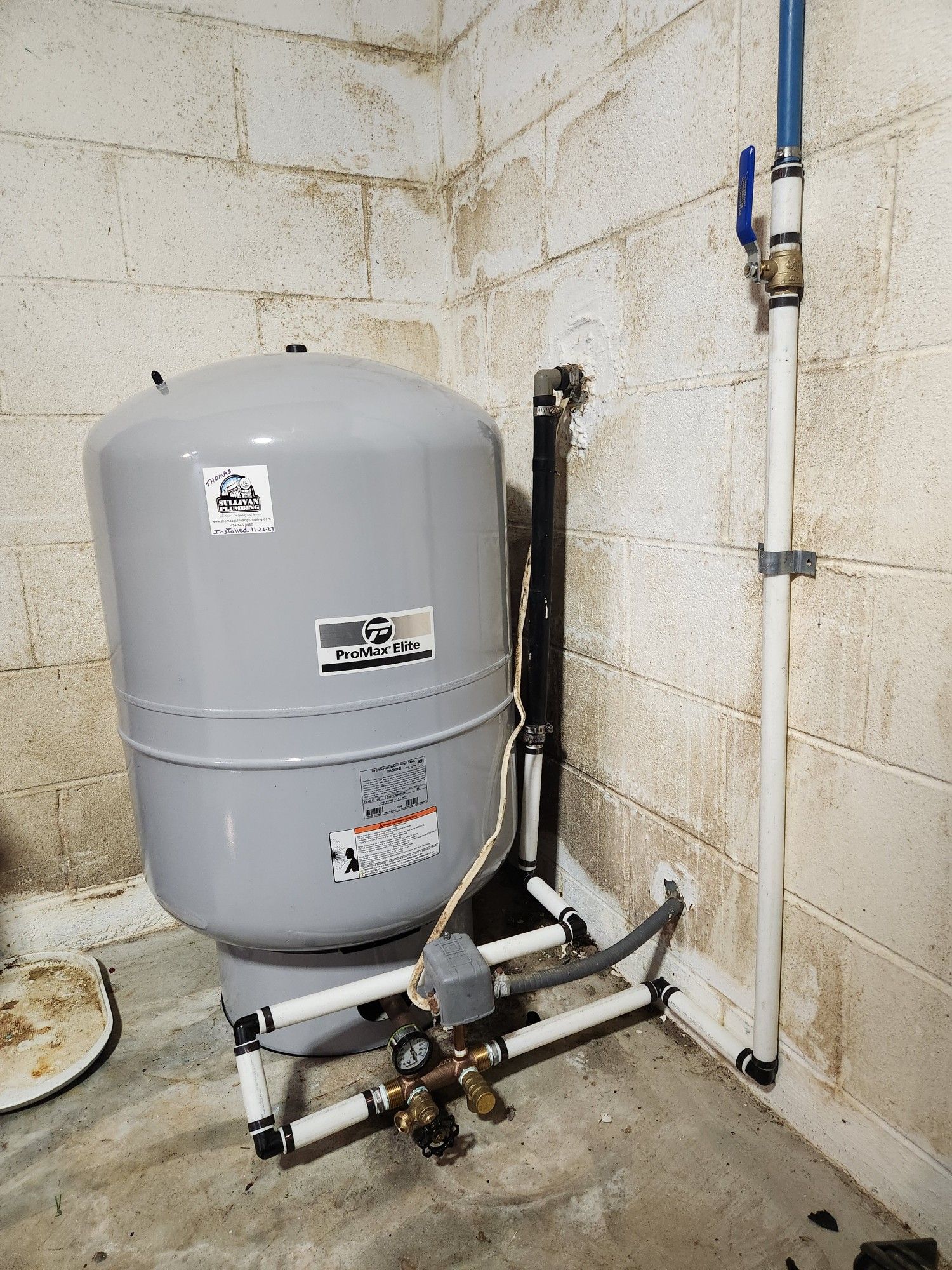 A water tank is sitting in a corner of a room next to a pipe.