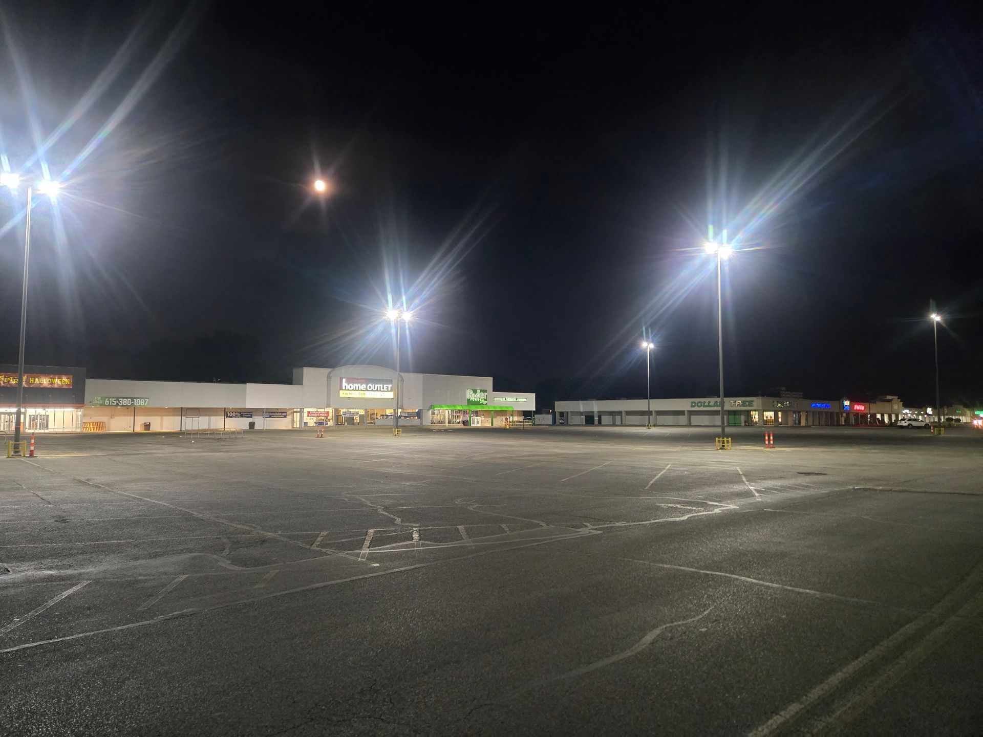 Empty parking lot at night with bright overhead lights illuminating a strip mall.