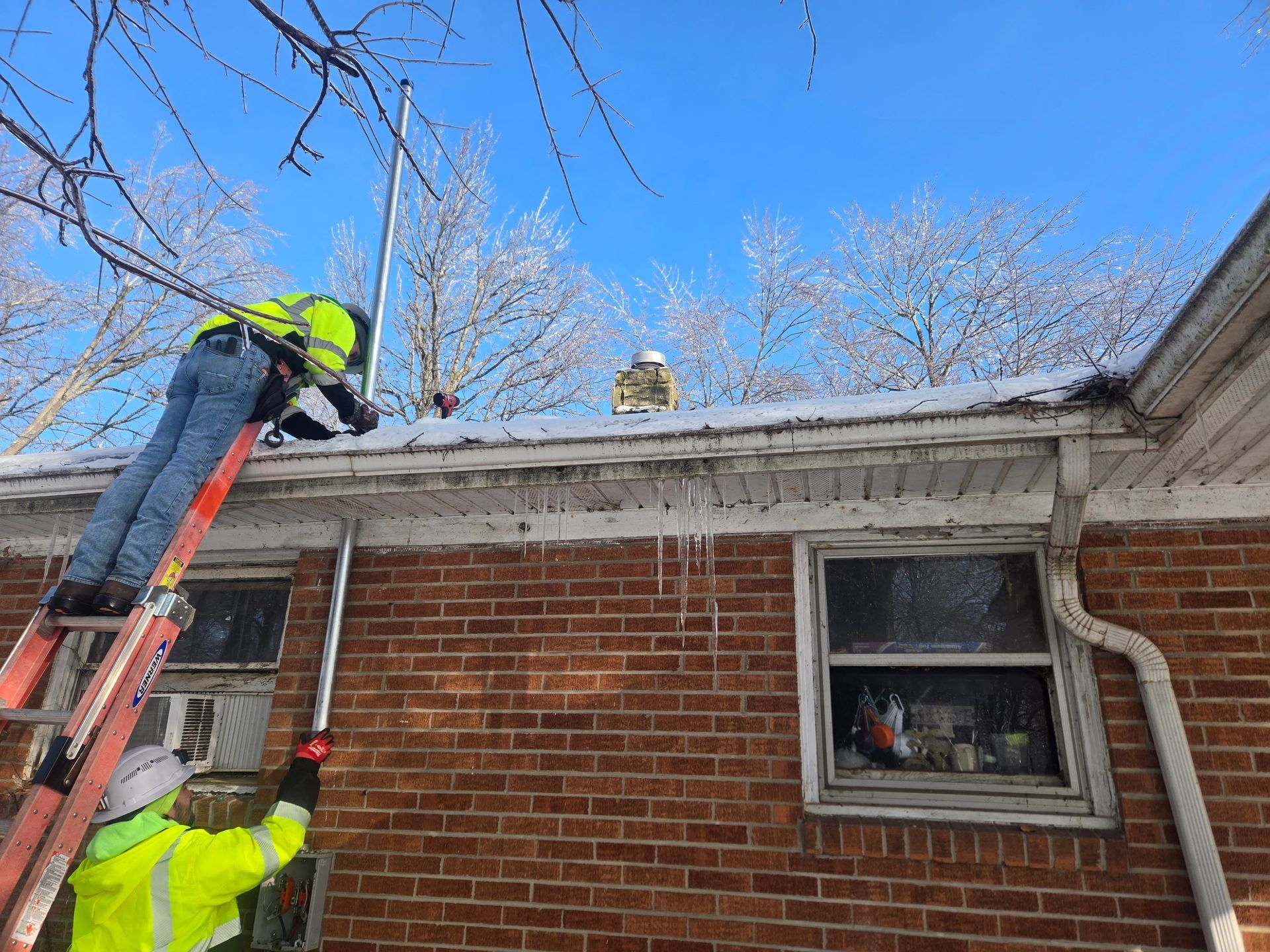 Two workers on a ladder, clearing ice from a roof's gutter. Cold, sunny day, brick house.