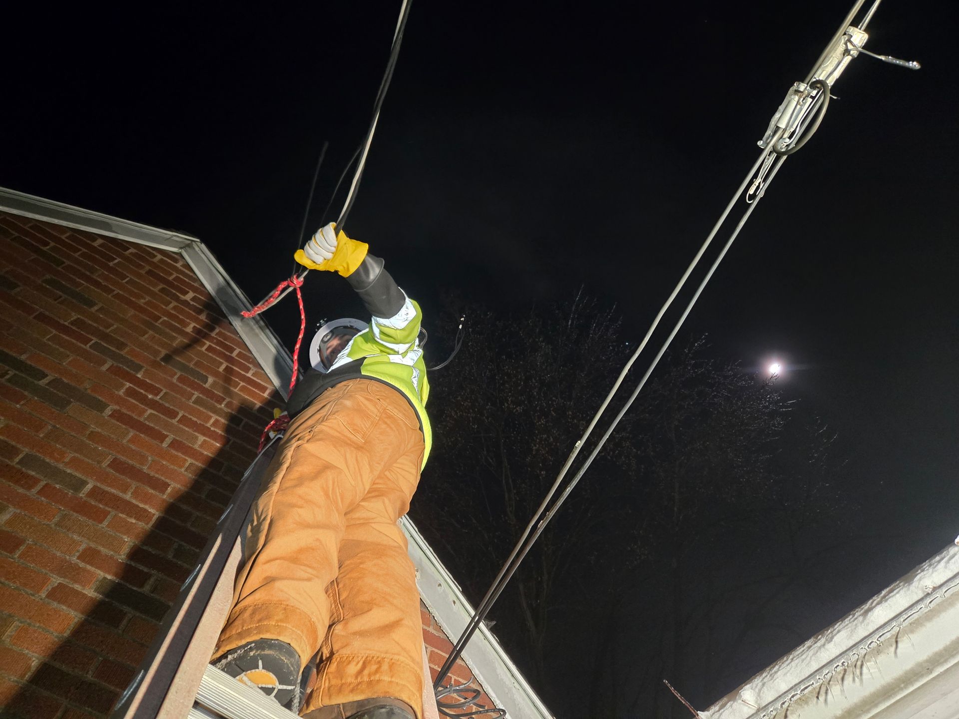 Lineman repairs power lines at night; he's on a ladder next to a brick building.
