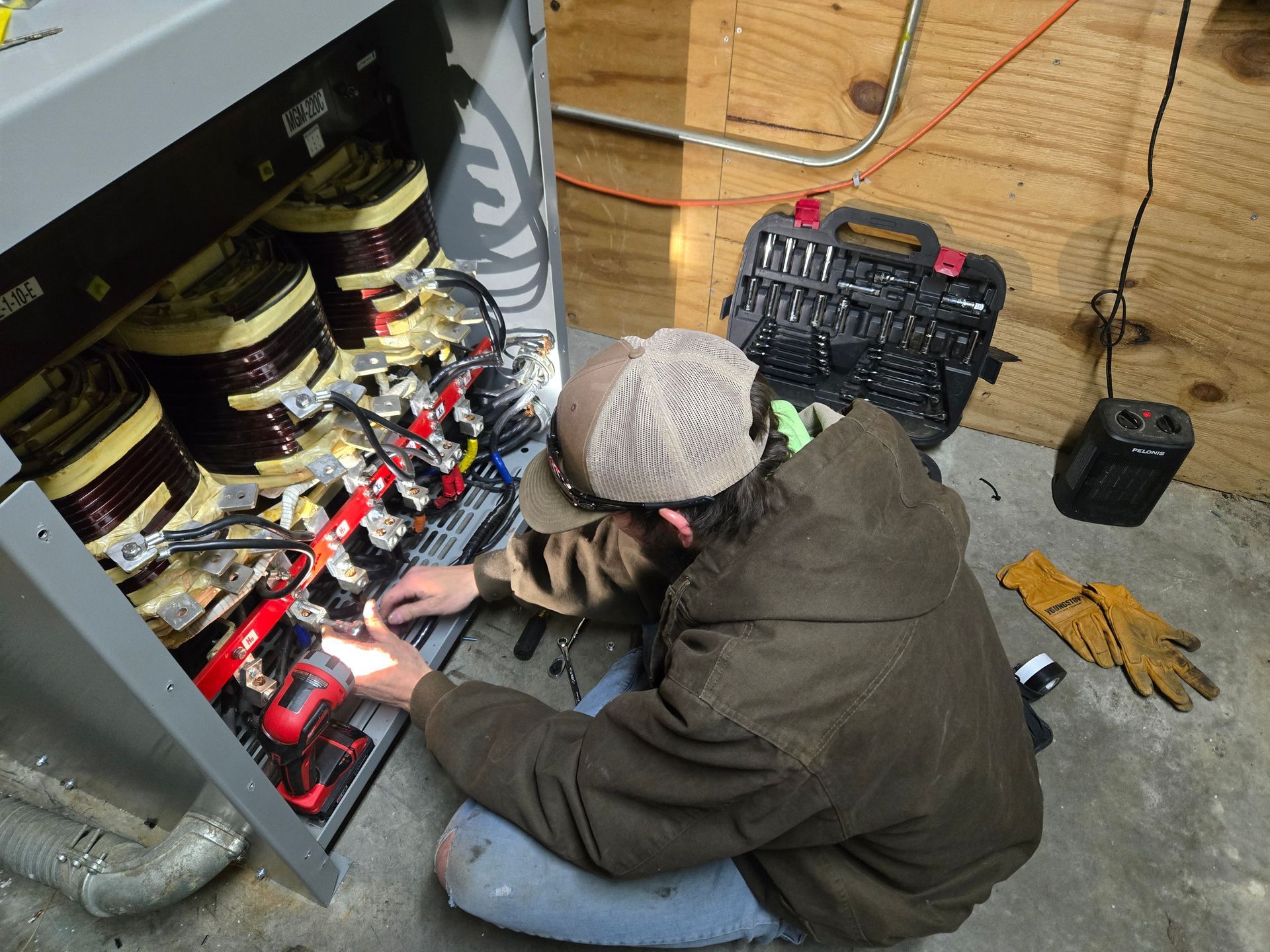 Person in a jacket working on electrical wiring inside a large cabinet. Tools and gloves are nearby.