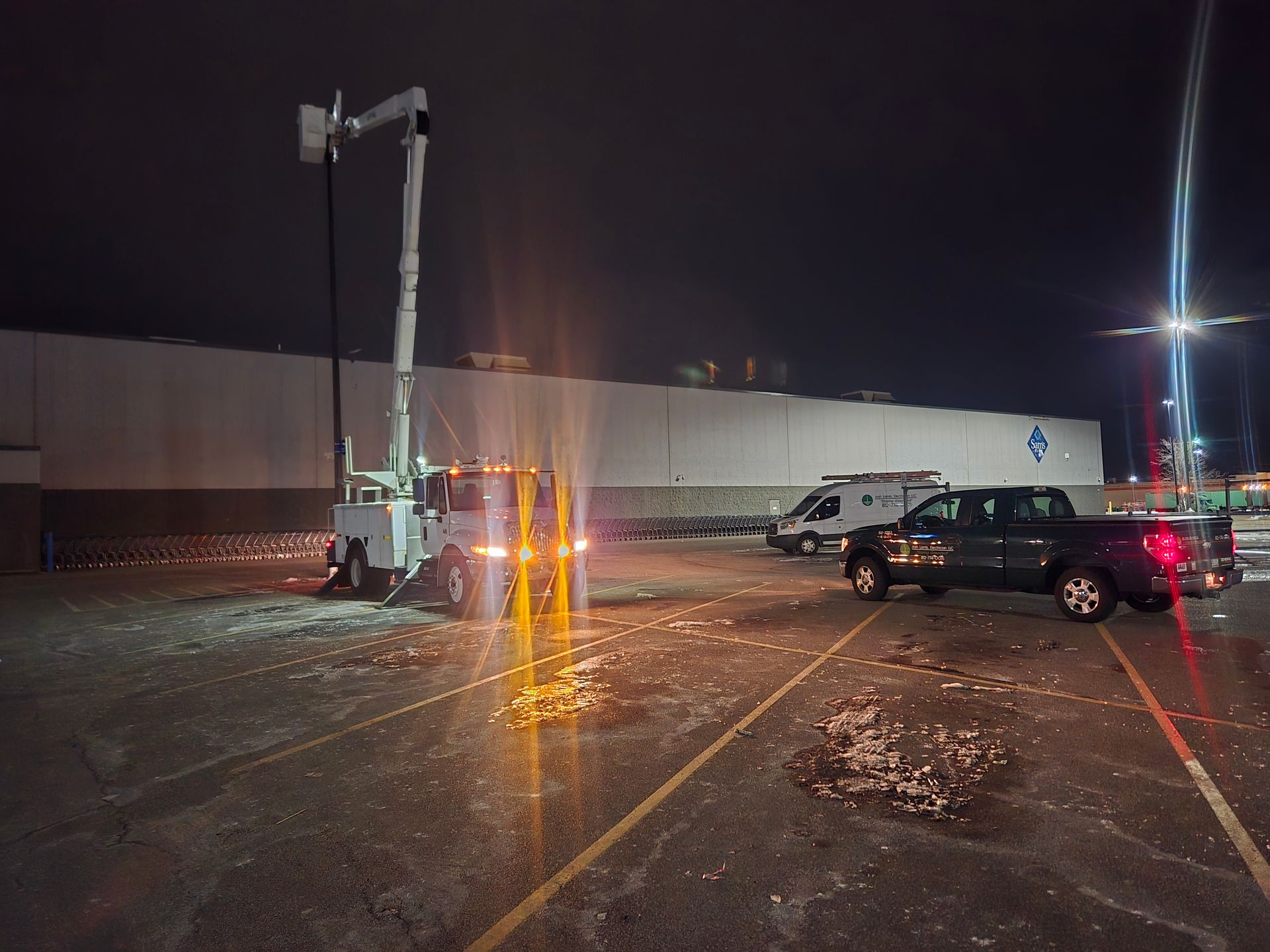 A bucket truck and work vehicles in a dark parking lot at night; the building in the background has lights.