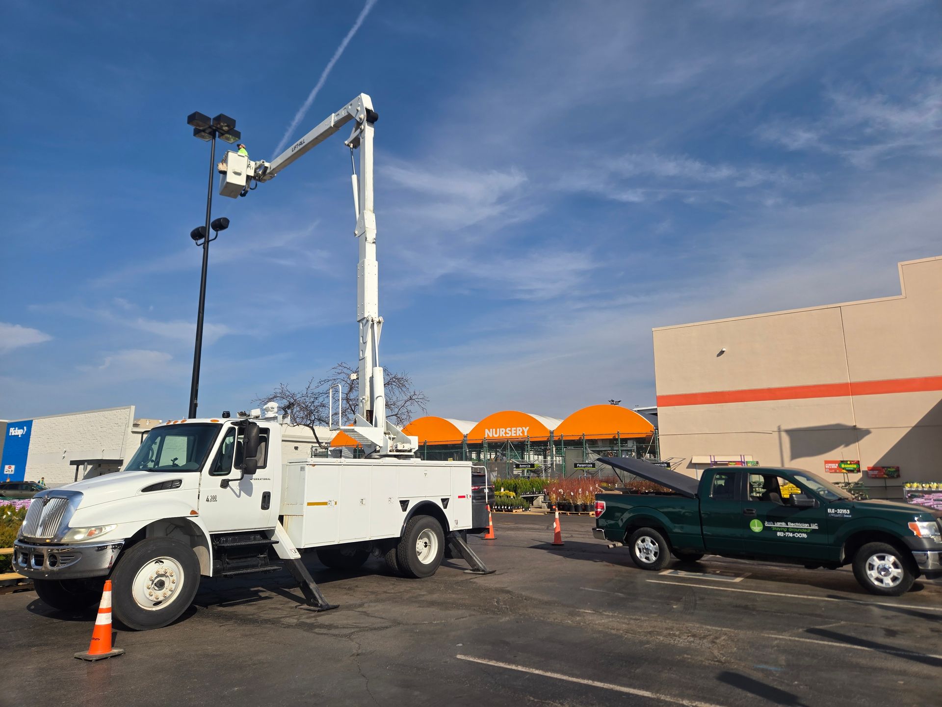White truck with extended arm near a Home Depot building; another green truck is also present, under a cloudy sky.