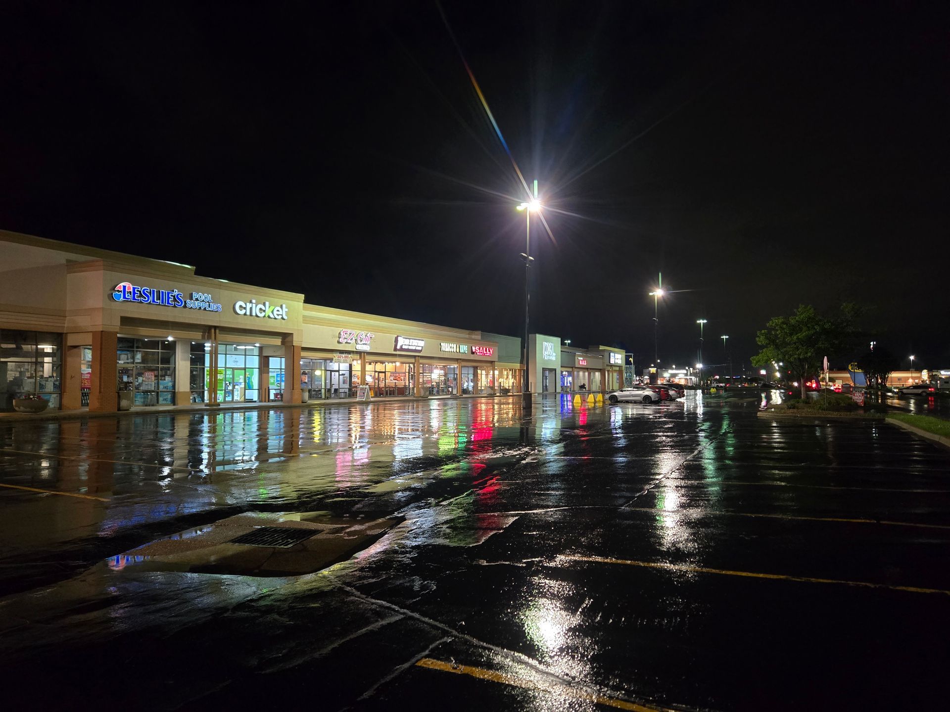 Night view of a shopping center parking lot after rain, reflecting bright store lights, with dark sky overhead.