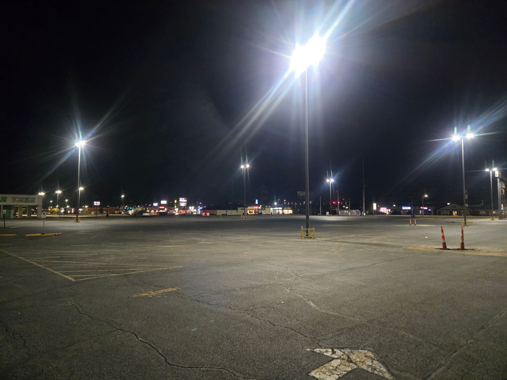 Empty parking lot at night, illuminated by bright street lights; distant store signs visible.