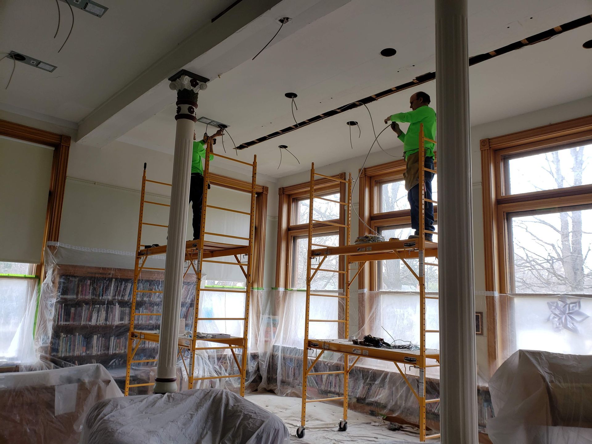Two workers on scaffolding install lights in a room with covered windows and furniture.